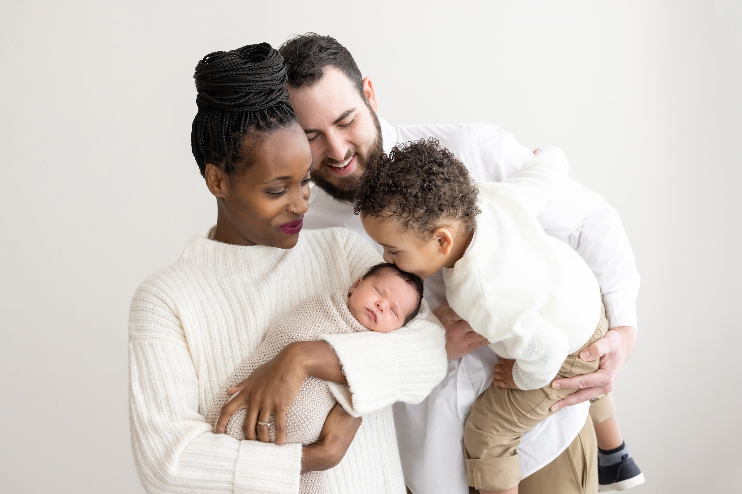  Family newborn portrait of a mother cradling her newborn in her arm as dad stands behind her holding their toddler son and the son is kissing his sister’s head taken in a studio near Buckhead in Atlanta, Georgia utilizing natural light. 