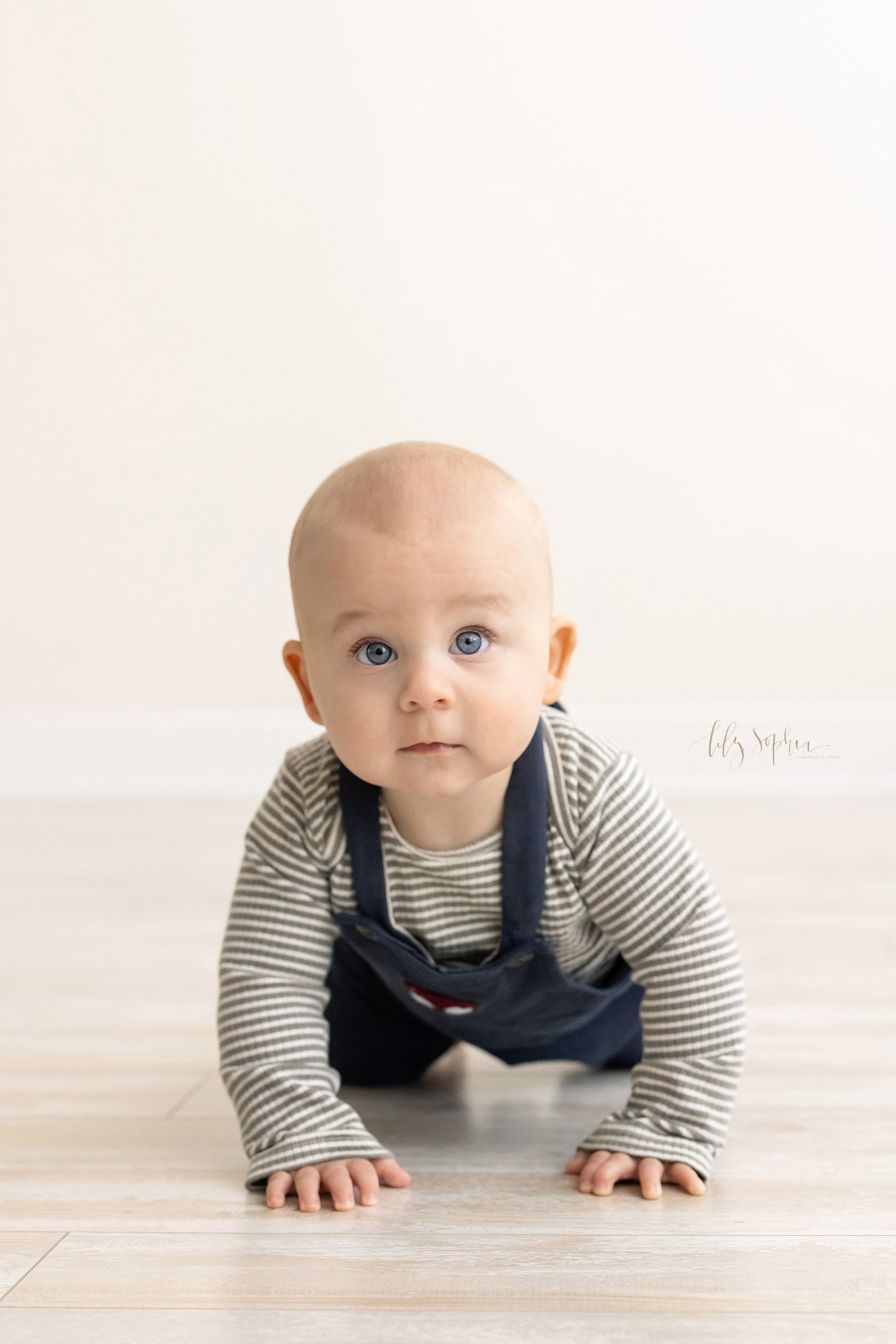  Milestone baby portrait of a six month old baby boy with huge blue eyes wearing a striped long-sleeved shirt and overalls crawls on the floor of a studio near Oakhurst in Atlanta that uses natural light. 