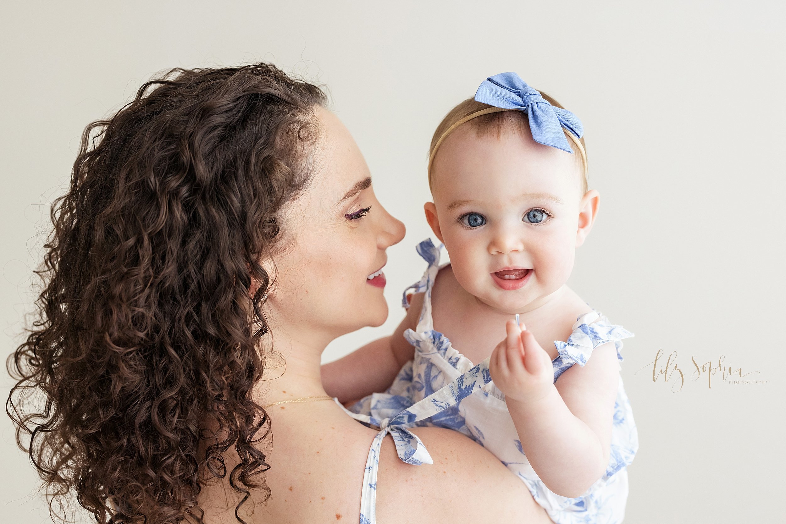  Mother and daughter photograph of a baby girl wearing a headband with a blue bow on it in her hair and mom and daughter wearing matching blue and white sundresses as mom holds her baby girl in her arms and looks at her while she peeks over her shoul