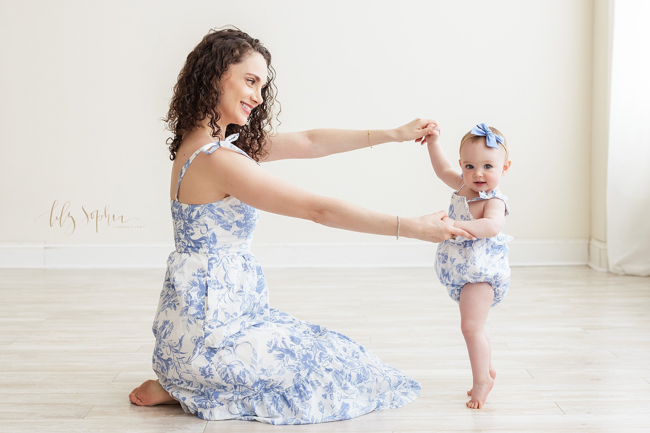  Family photo session of a mother and daughter with mom sitting on the floor of a studio holding the hands of her baby girl as her daughter stands on her tiptoes in front of a window streaming natural light in a studio near Poncey Highlands in Atlant