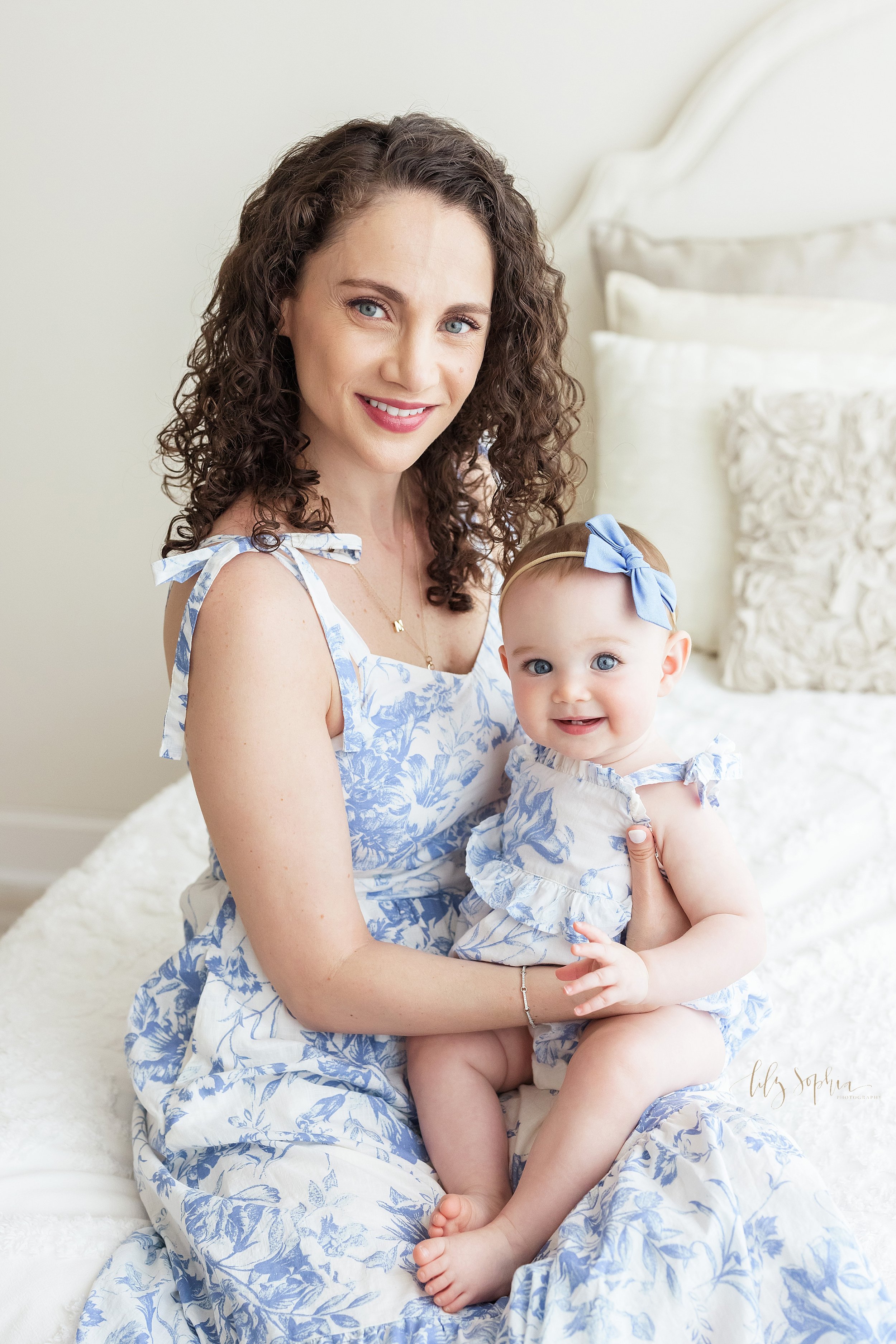  Family photo session with a mother and her baby as mom sits on a bed in a natural light studio wearing a blue and white sundress and her daughter sits on her lap wearing a sun suit in the same blue and white fabric taken near Kirkwood in Atlanta. 