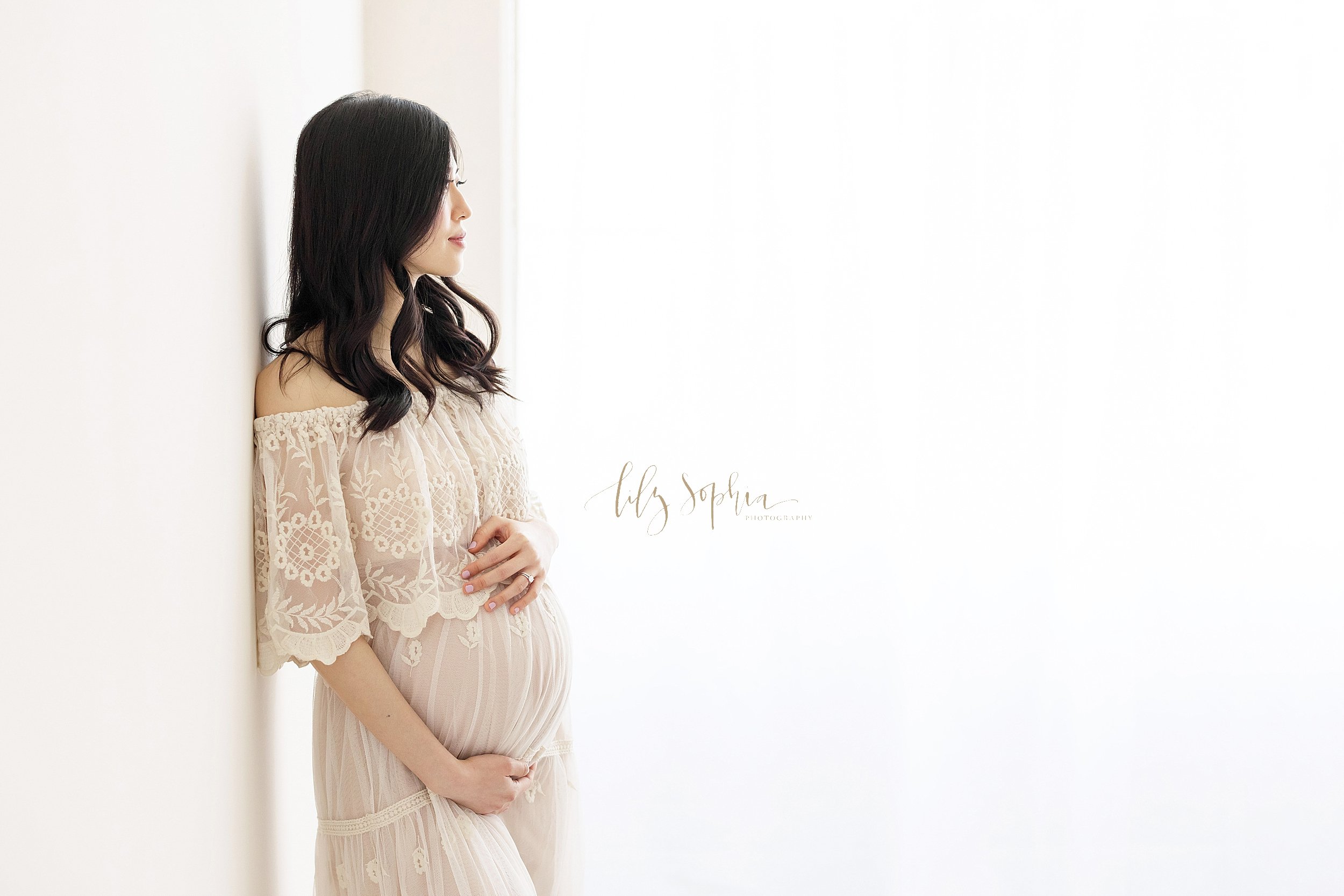  Maternity photograph of an Asian woman with her right shoulder against a wall as she wears a full-length flowing off-the-shoulder gown with a large lace ruffle bodice with her hands framing her belly as she contemplates the birth of her child while 