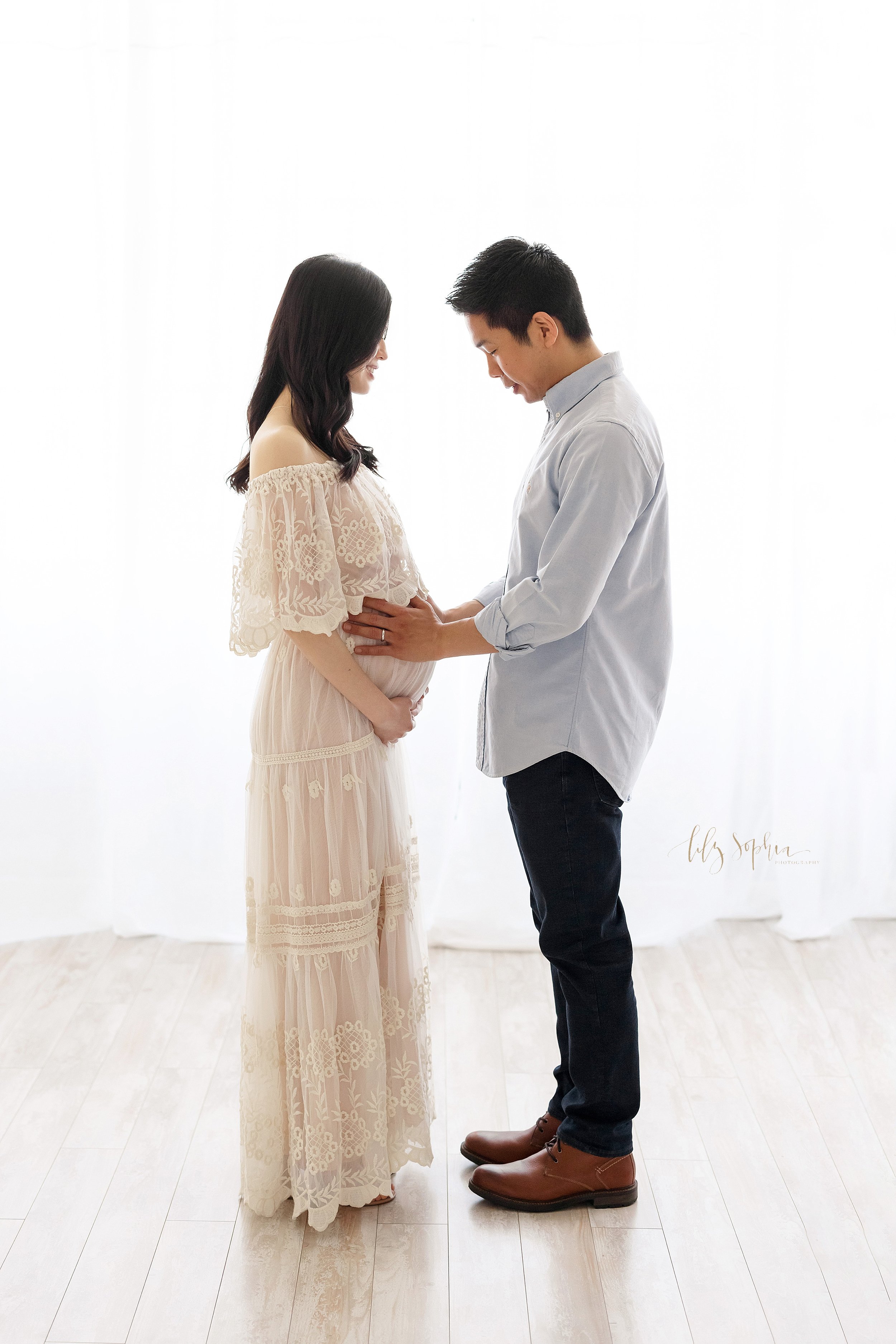  Maternity photo session of an Asian couple with the wife wearing  an off-the-shoulder full-length gown with a lace ruffle bodice as she stands facing her husband with her hands at the base of her belly and her husband with his hands on their child i