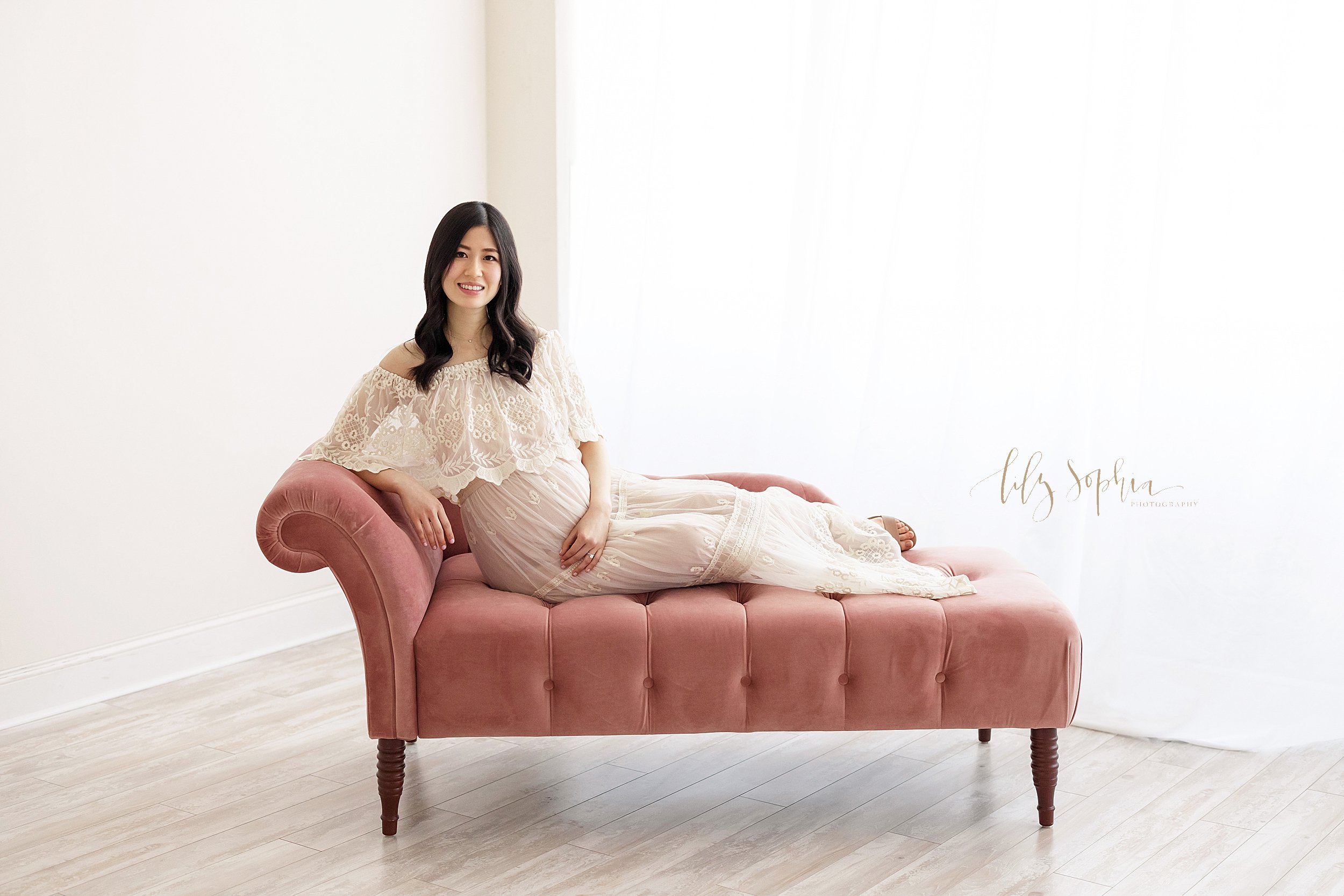  Maternity picture of an Asian woman as she lounges on a one arm tufted settee in front of a natural light window in a studio near Cummings in Atlanta. 