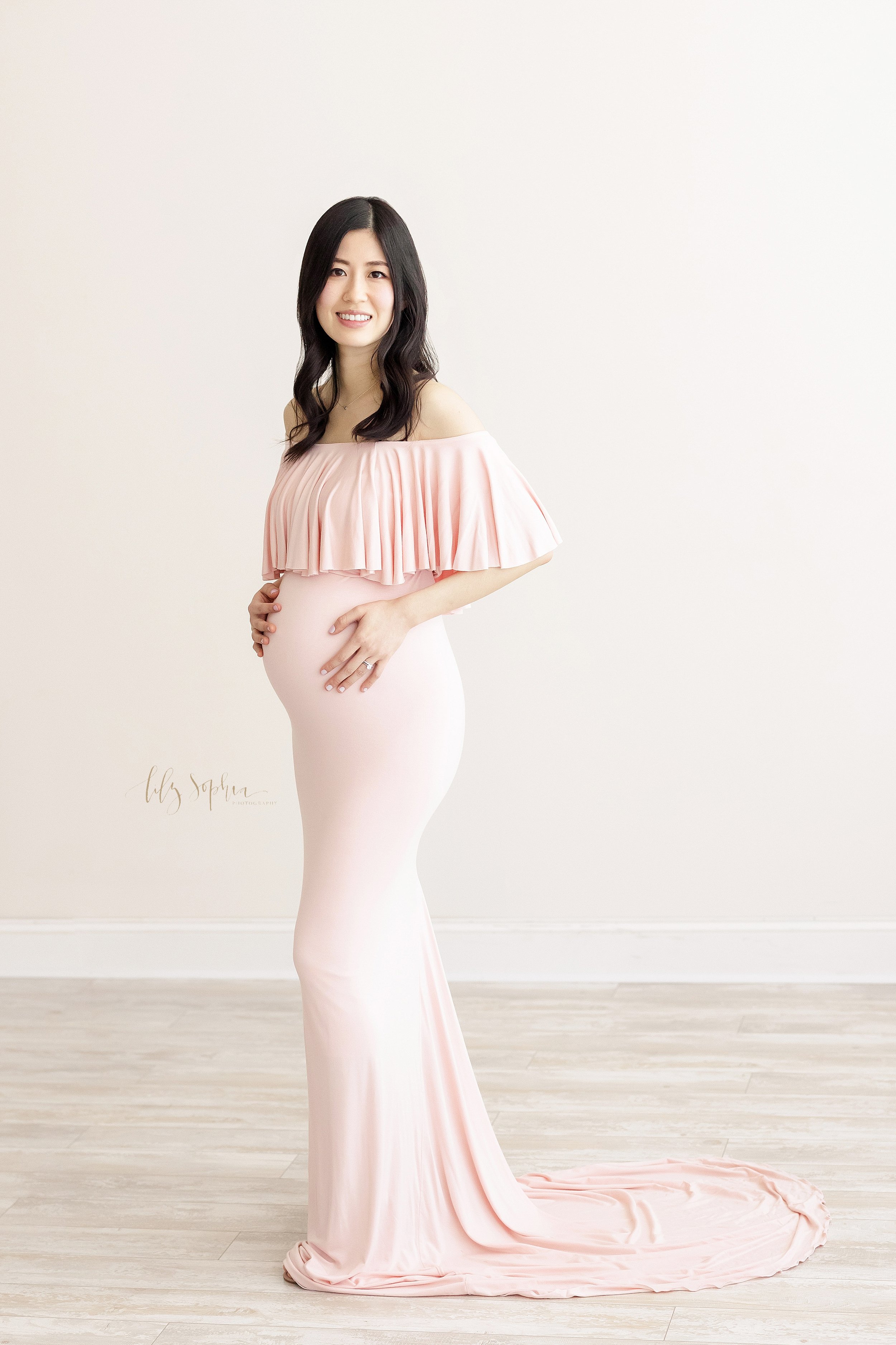  Maternity photo shoot with a pregnant Asian woman wearing a form fitting pink jersey off-the-shoulder gown with a ruffle bodice as she stands with her hands on her child in utero in a natural light studio near Midtown in Atlanta. 
