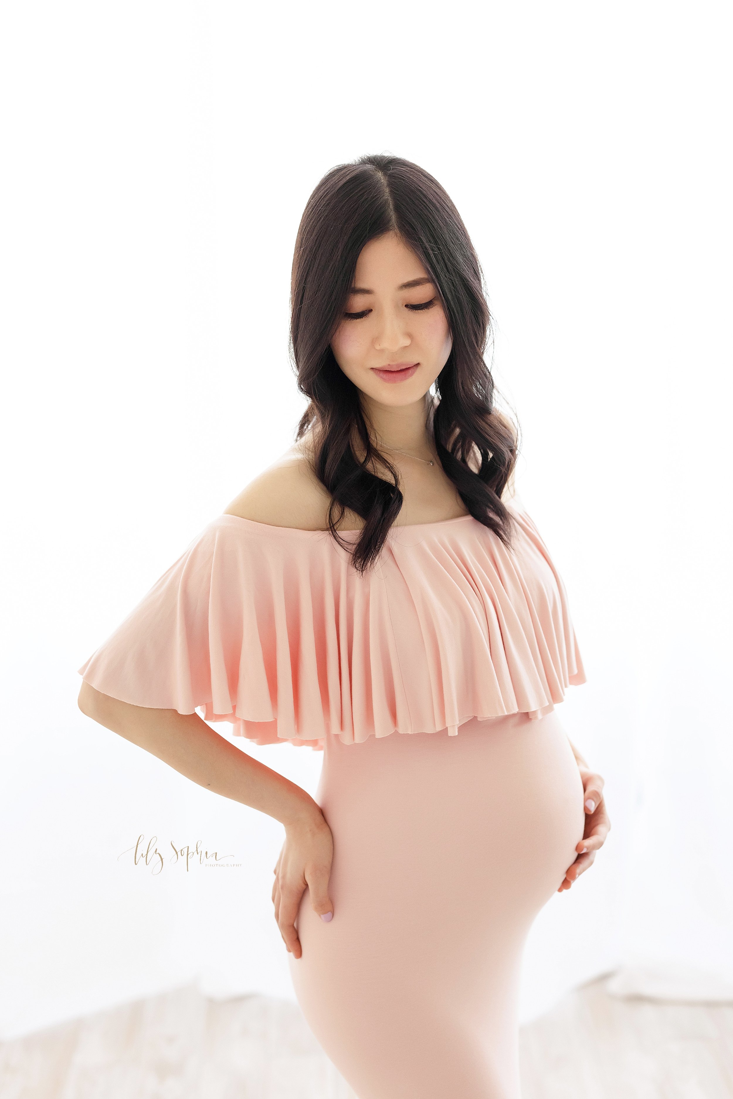  Close-up maternity portrait of an Asian pregnant woman wearing a pink form-fitting jersey off-the-shoulder gown with a large pink ruffle bodice as she stands in front of a window streaming natural light with her right hand on her hip and her left ha