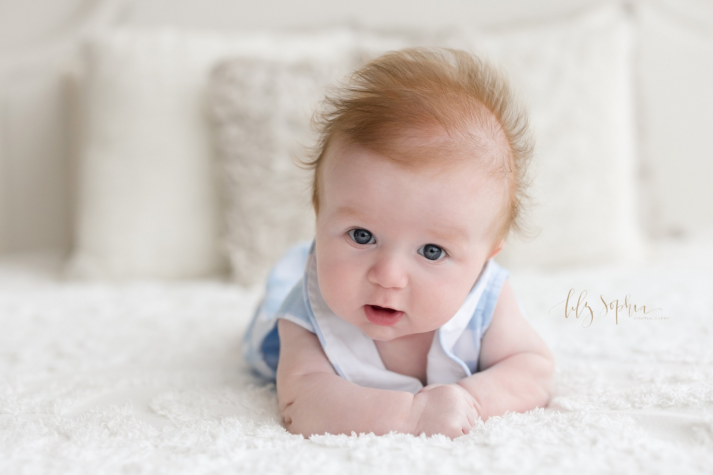  Baby portrait of a wispy haired red-headed three month old little boy as he lies on his stomach on a bed and lifts his head to show his big blue eyes taken in a natural light studio near Midtown in Atlanta. 