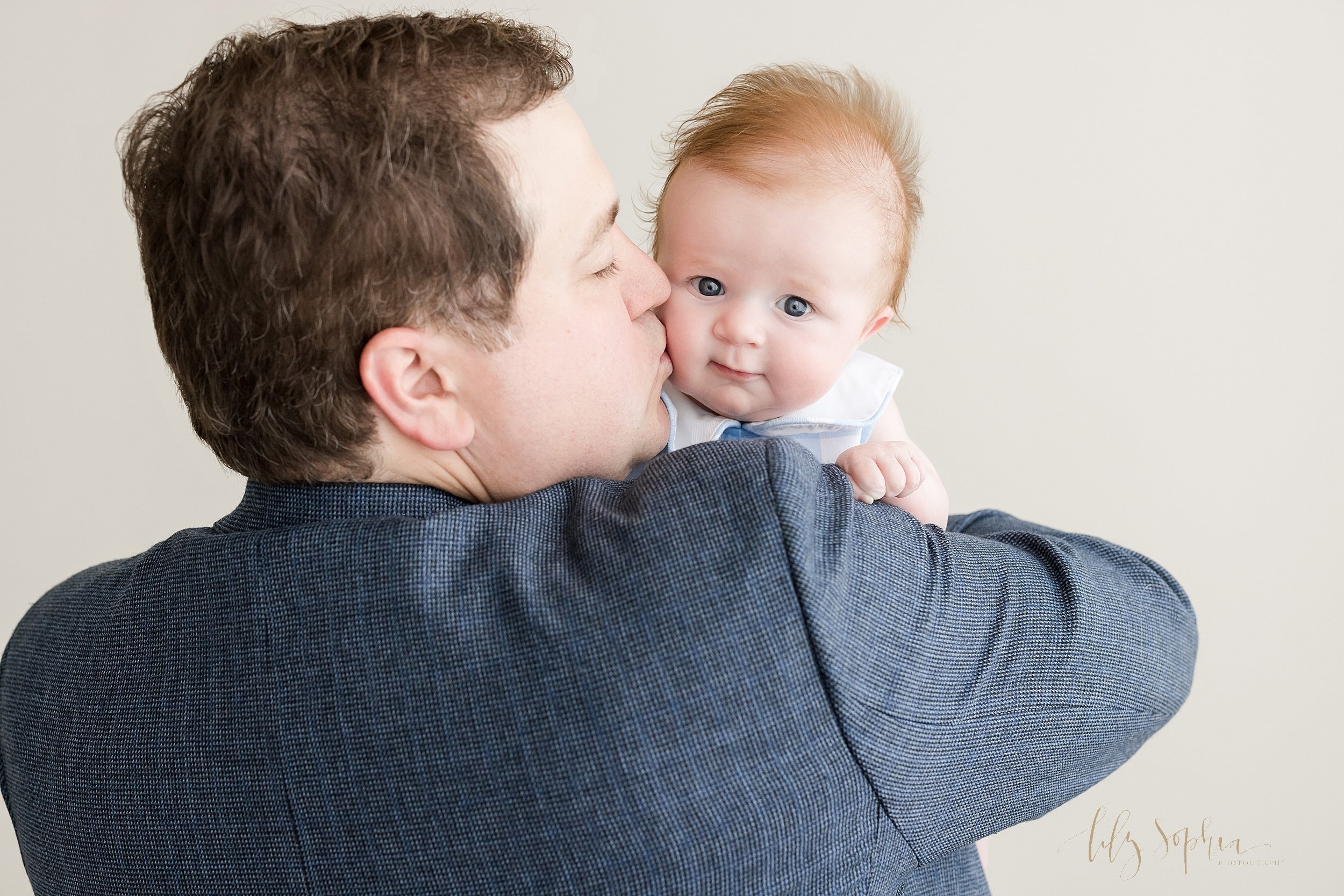  Family photo of a three month old baby boy peeking over his father’s shoulder as his father holds him and kisses him on his cheek taken near Decatur in Atlanta in a natural light studio. 