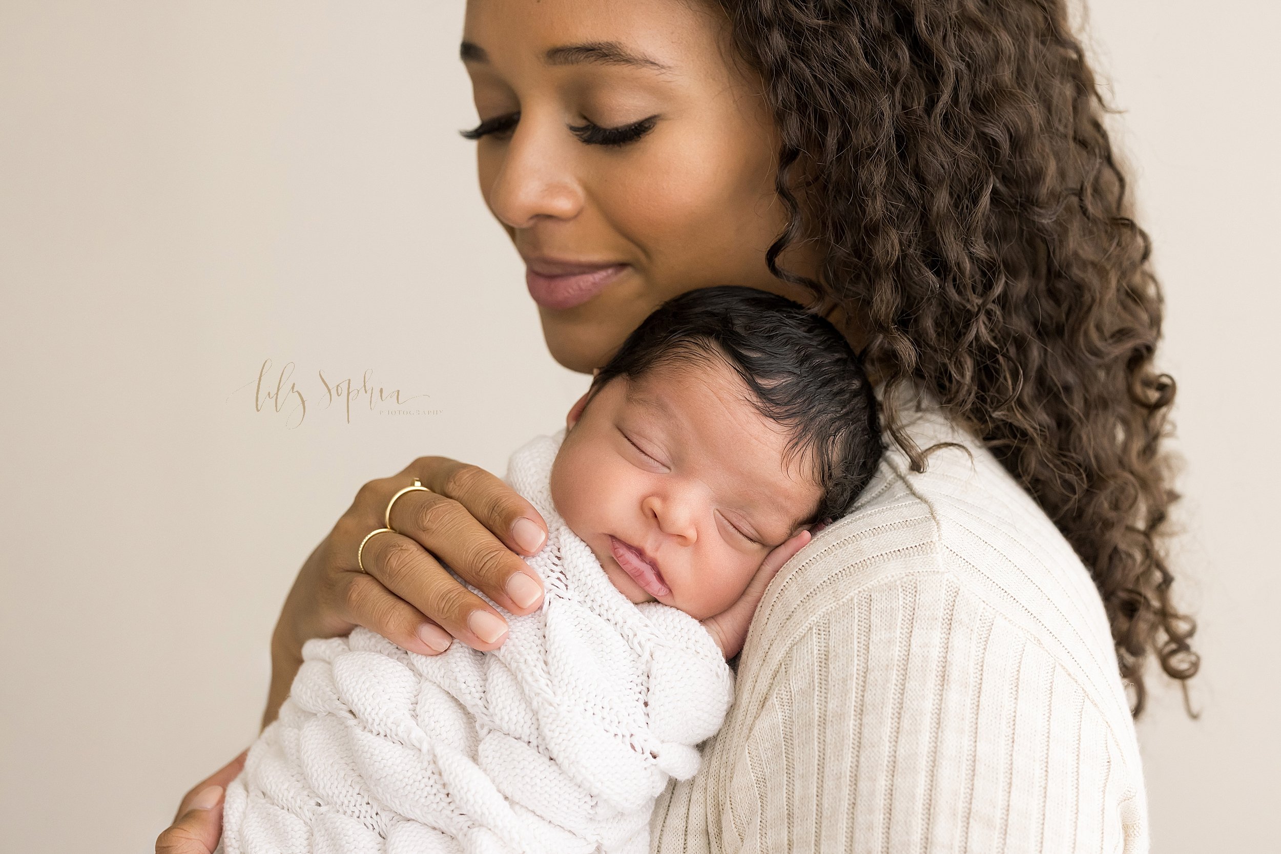  Newborn photo shoot of an African-American infant girl wrapped in a soft white blanket as she sleeps with her tiny hand under her head against her mother’s shoulder as her mother stands in front of a window streaming natural light in a studio near D