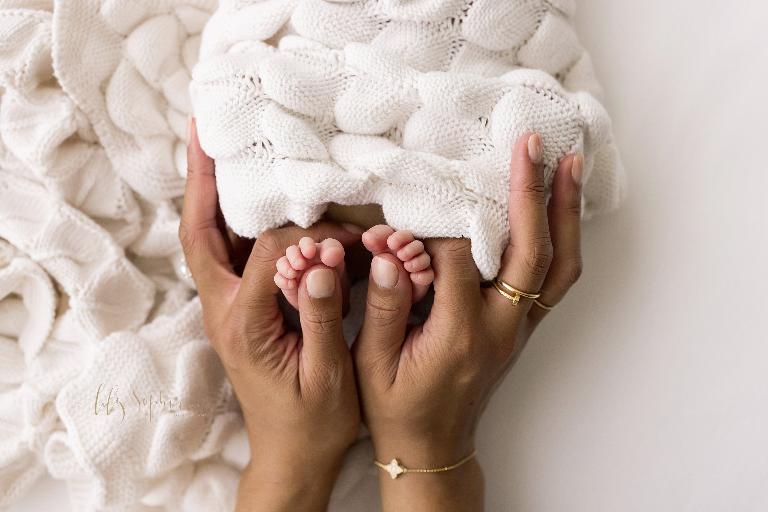  Newborn photo session of an African-American infant girl with her tiny feet peeking out from under a soft white crocheted blanket being held by her mother taken in a natural light studio near Morningside in Atlanta. 