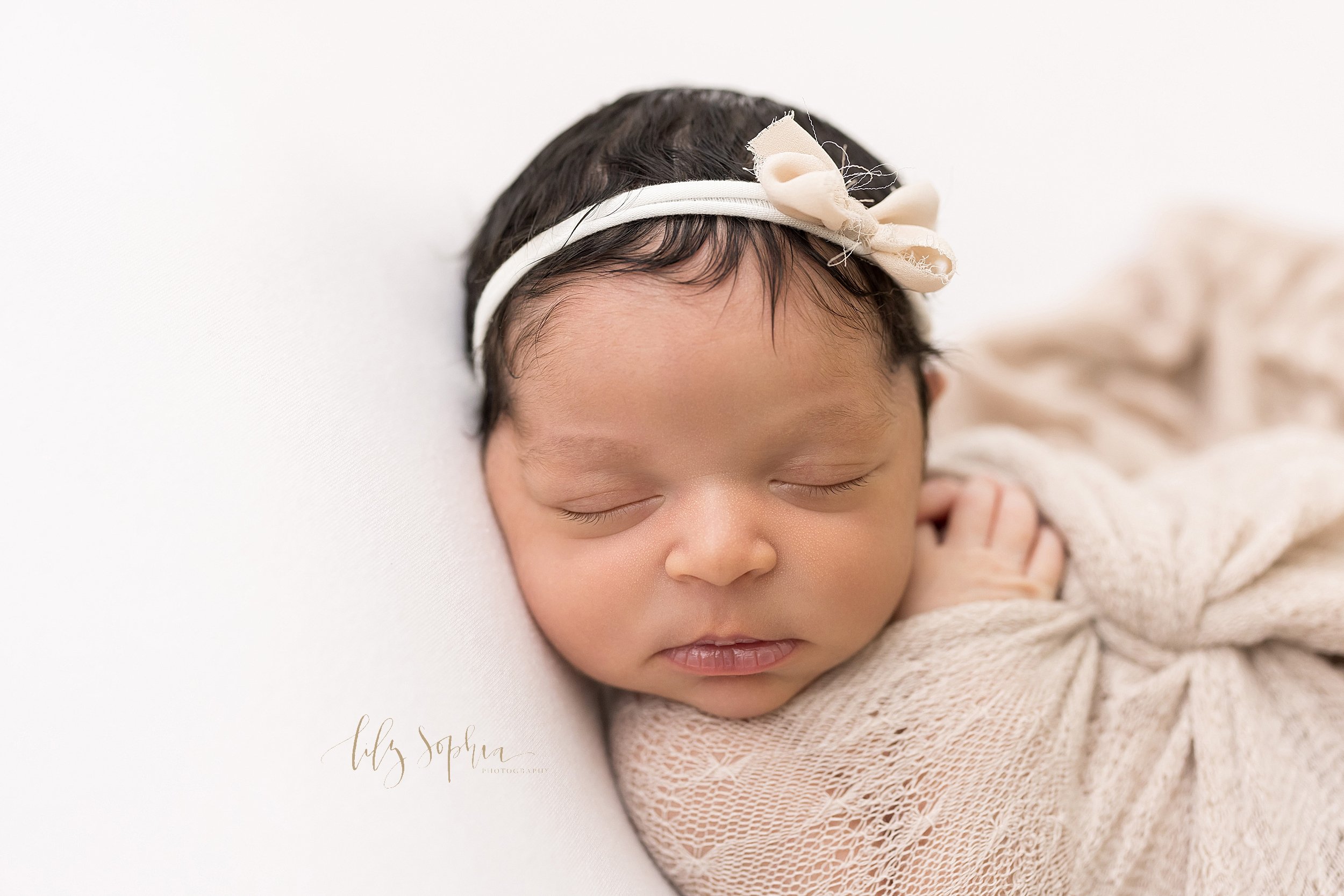  Newborn close-up photo of the face of an African-American infant girl wearing a headband with a bow on her head as she peacefully sleeps with her hands by her neck while wrapped in a blanket and lying on her back taken near Kirkwood in Atlanta in th