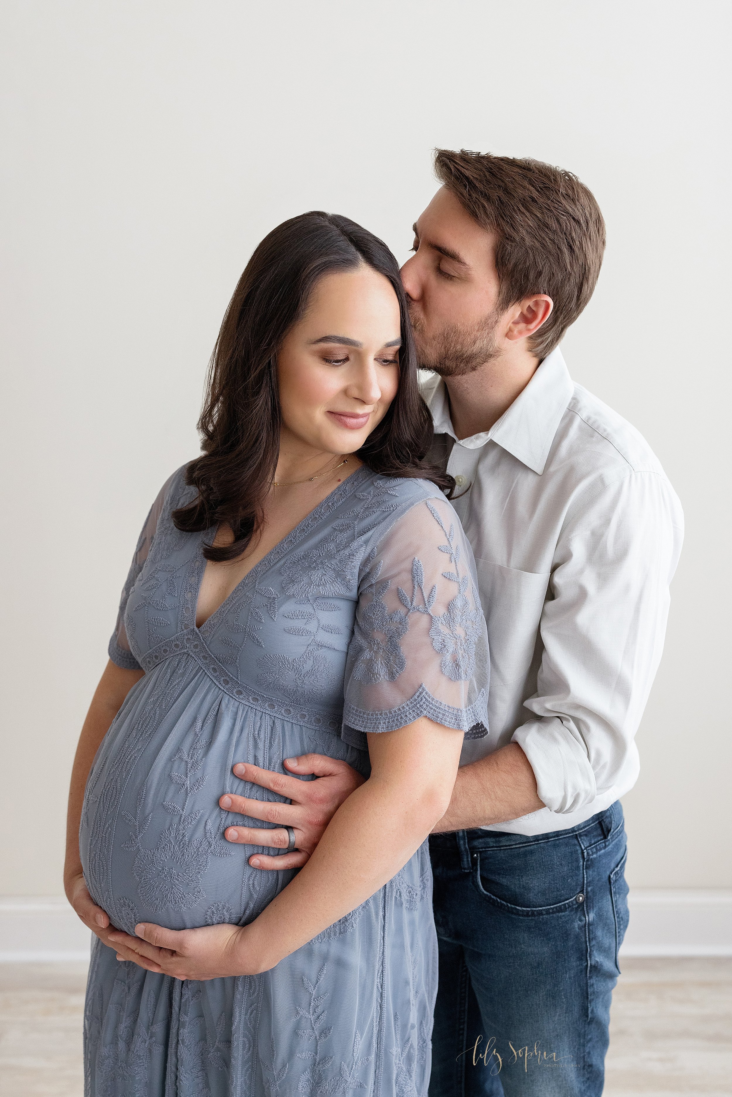  Maternity photo of an expectant mother looking over her right shoulder as she holds her belly with her hands and her husband stands behind her with his hands on her belly and kisses her head taken in a natural light studio near Old Fourth Ward in At