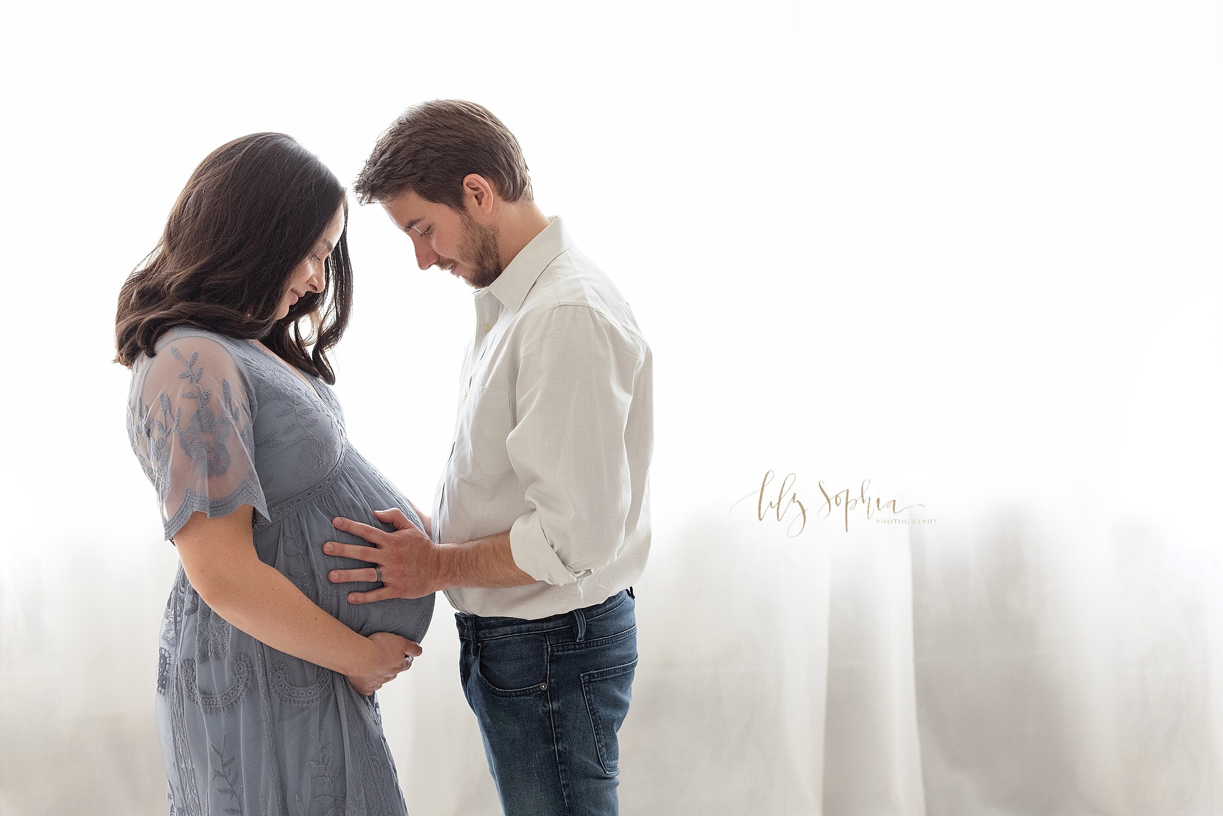  Maternity session with a pregnant mother standing facing her husband in front of a window streaming natural light with the husband placing his hands on his child in utero and his wife framing her belly taken in a studio near Midtown in Atlanta, Geor