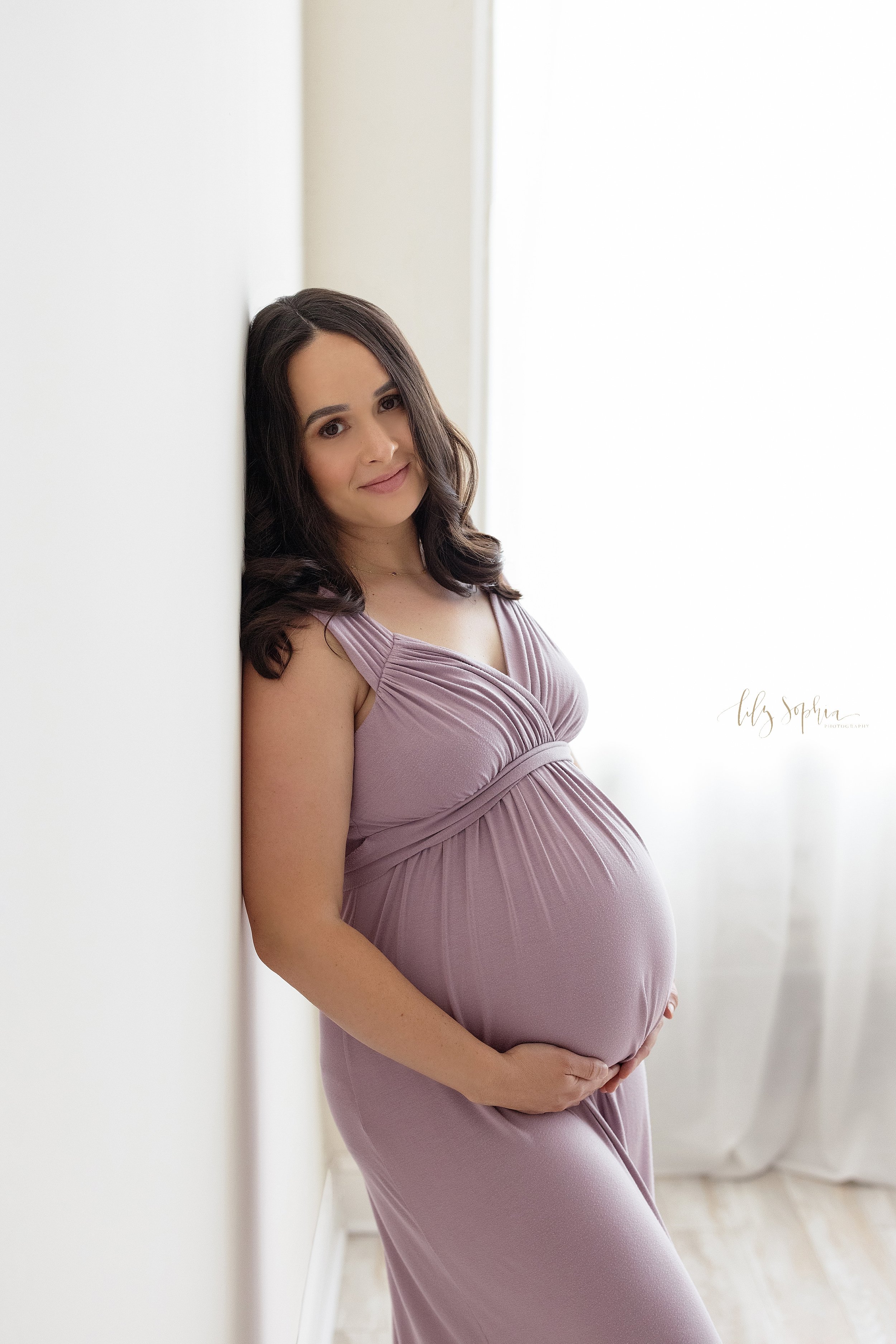  Maternity session with an expectant mother wearing a soft jersey knit gown as she stands with her back against a wall in front of a window streaming natural light and frames her belly with her hands taken in a studio in Ponce City Market in Atlanta.