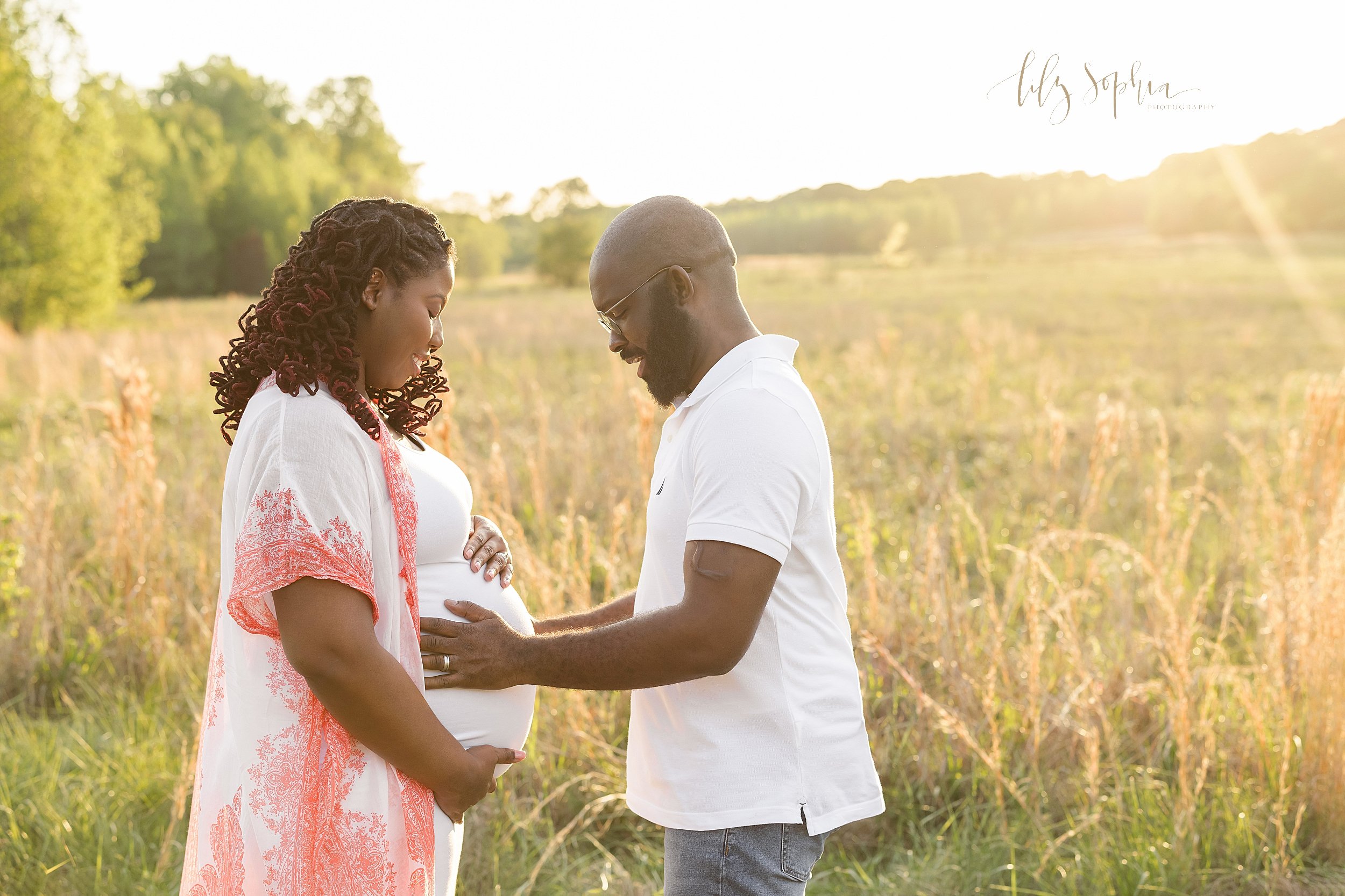  Maternity photo shoot with an expectant African-American couple as they face one another and the mother frames her belly with her hands and her husband places his hands on their child in utero while they stand at sunset in a field near Atlanta. 