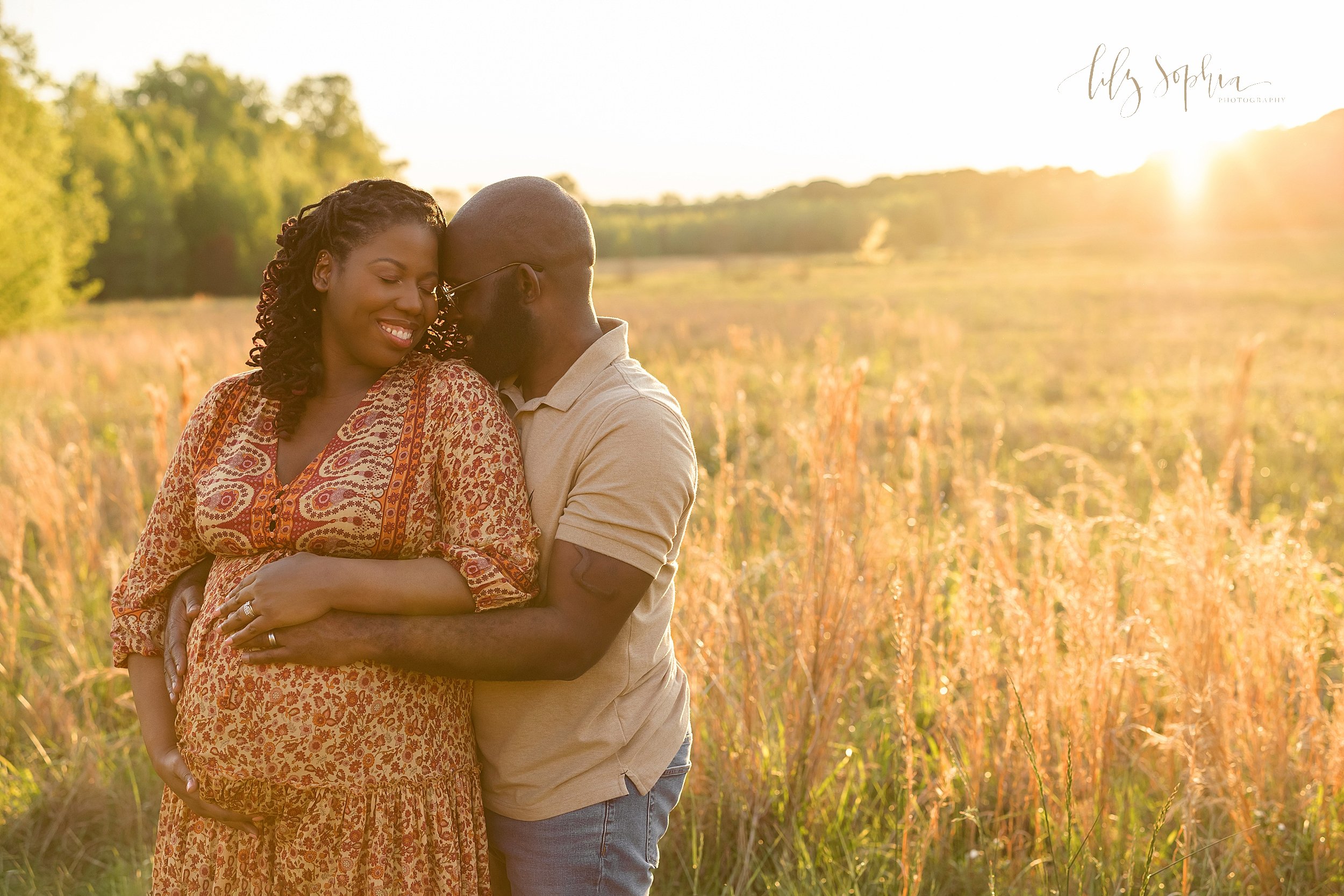  Maternity photo session of an African-American expectant mother as she stands in a field and frames her belly with her husband standing to her left side with his chin on her shoulder and holding his wife’s waist with his hand on their child in utero