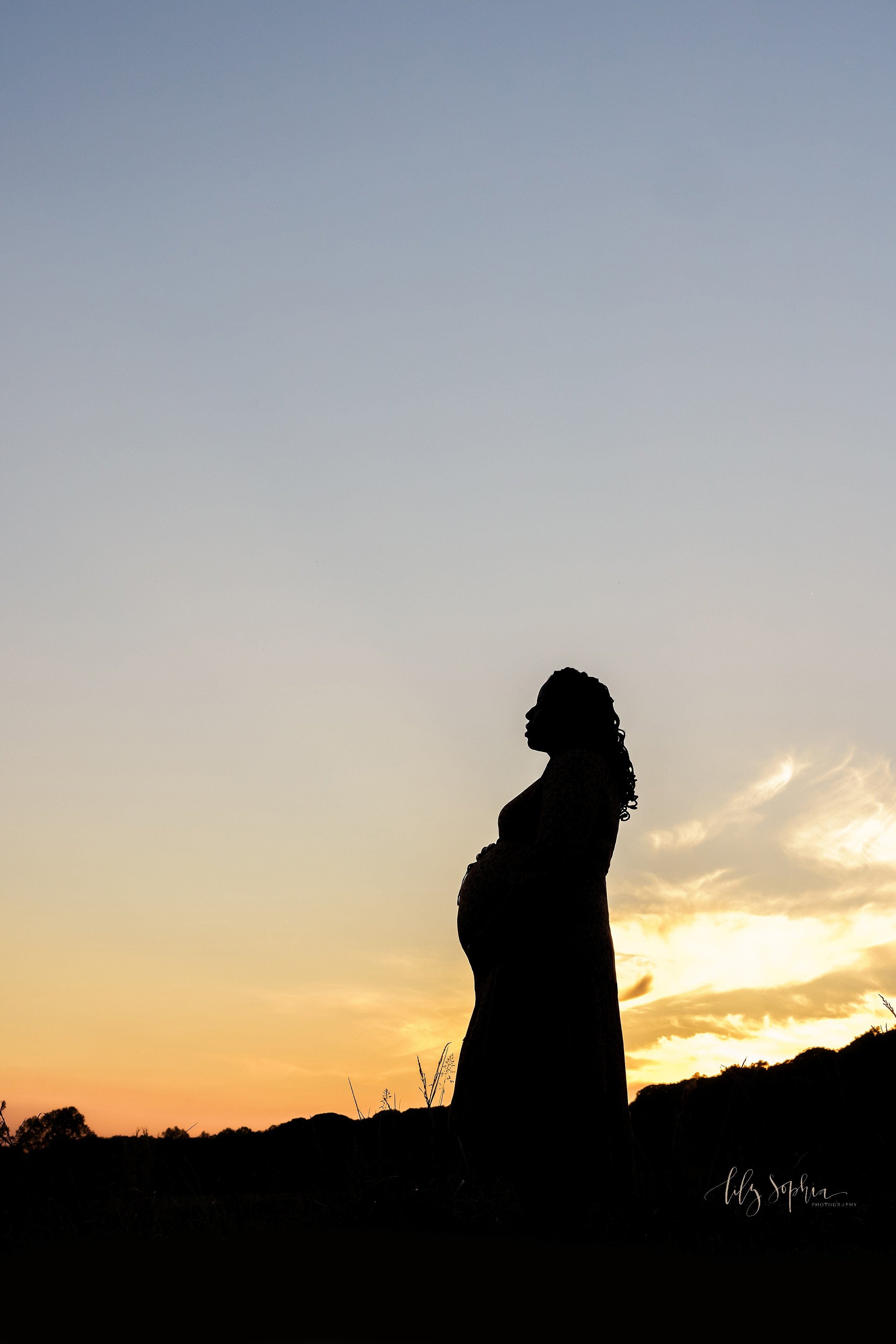  Silhouetted maternity portrait of an African-American mother as she stands atop a hill at sunset taken near Atlanta. 