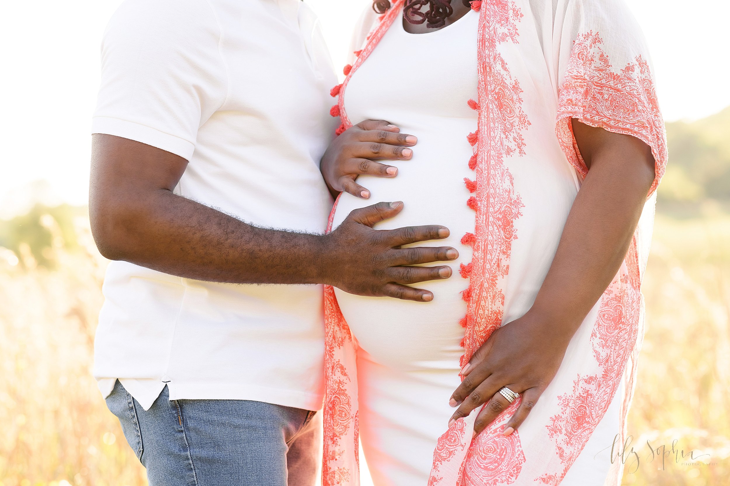  Close-up maternity photograph of an African-American woman’s belly as she and her husband place their hands on their child in utero taken near Atlanta in a field at sunset. 