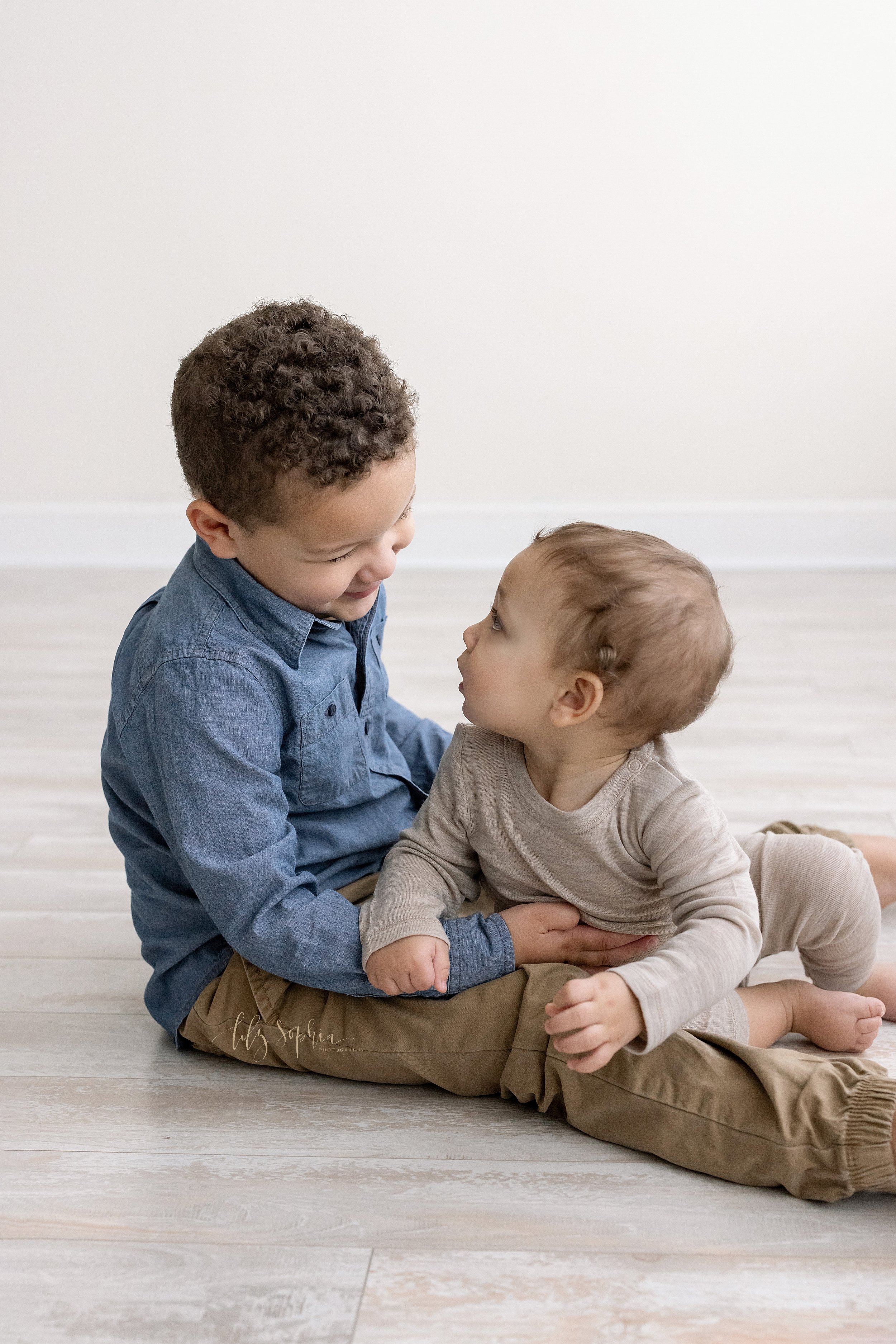  Family portrait of brothers as the older brother sits on the floor of a natural light studio with his seven month old baby brother sitting between his legs and the younger brother turns to face his brother and the two of them talk taken near Morning