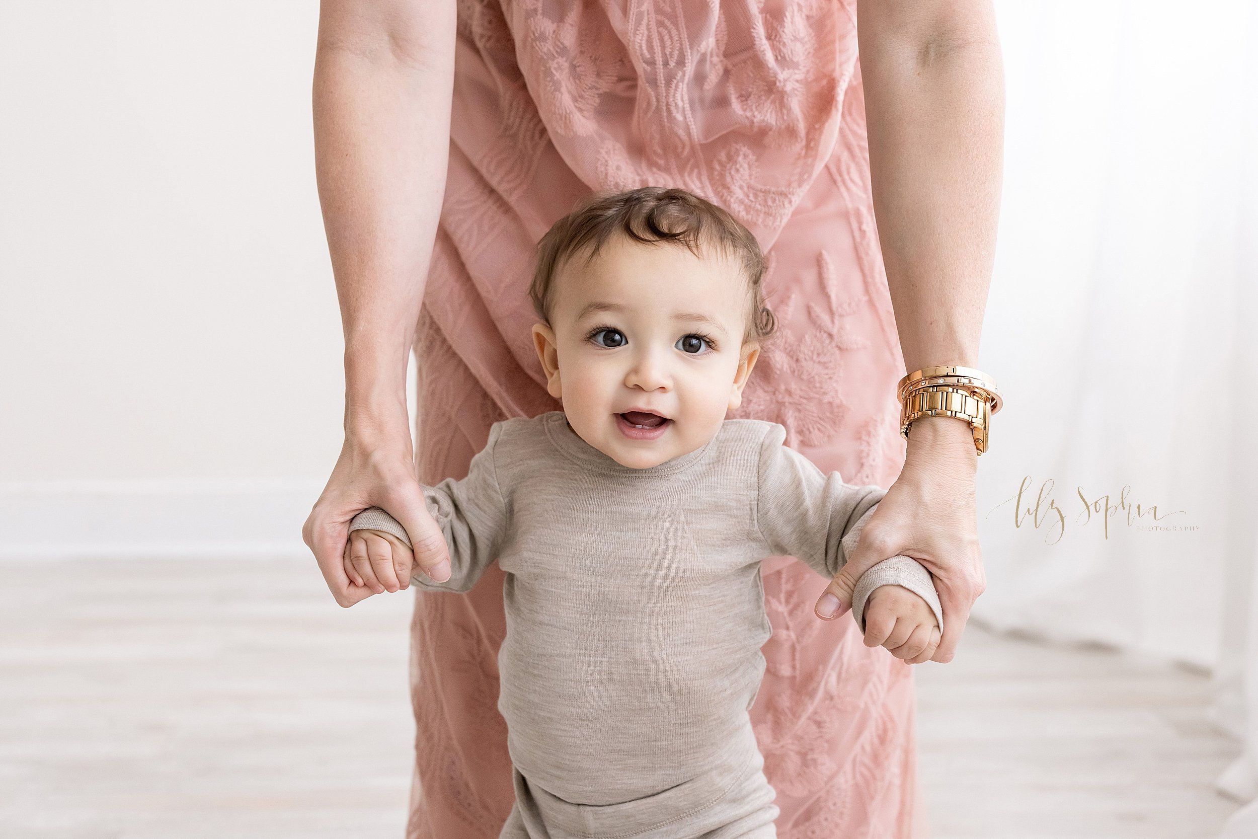  Family photo of a seven month old baby boy with his mother holding his arms as he walks in a natural light studio near Buckhead in Atlanta. 