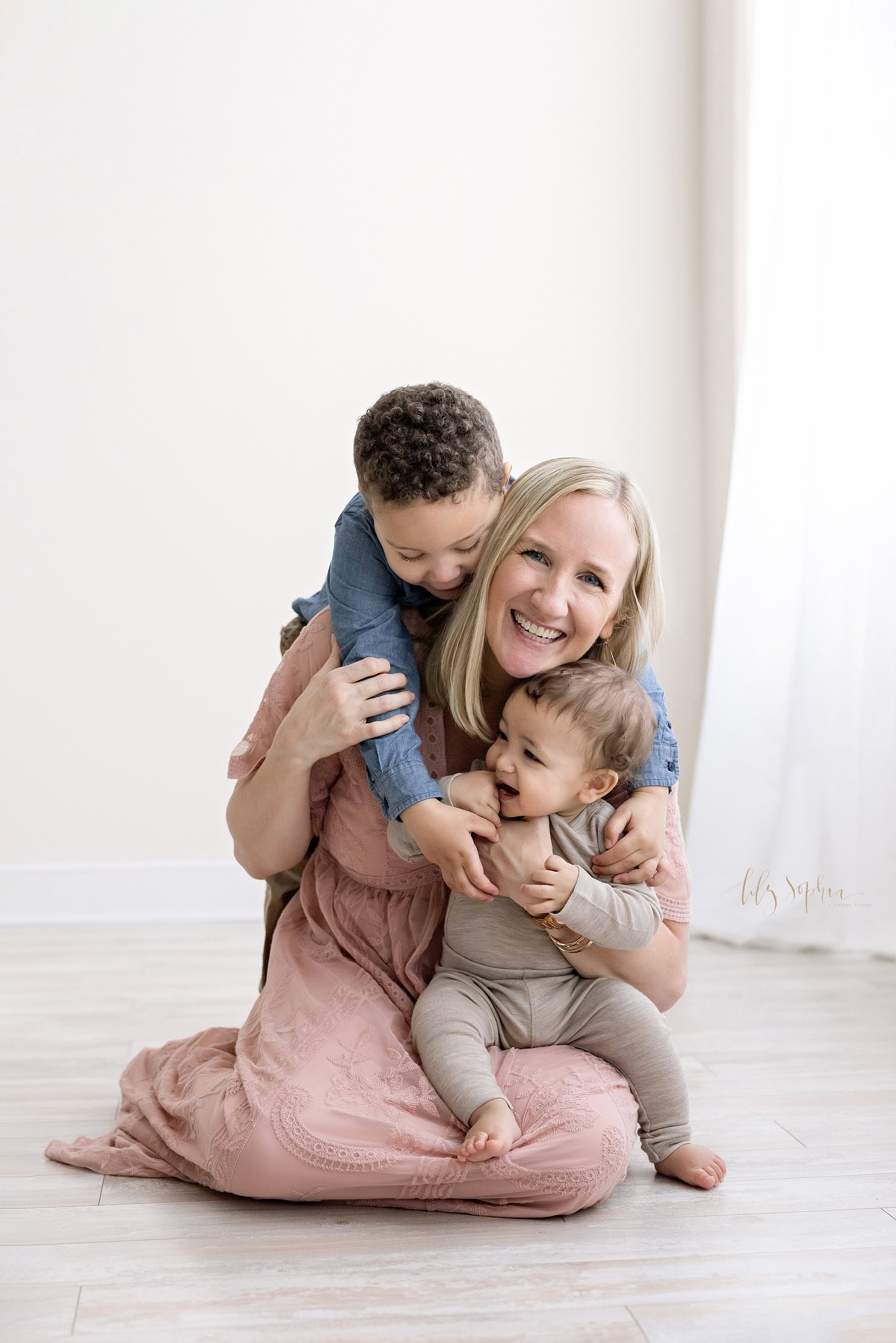  Fun family photo of a mother sitting on the floor of a studio in front of a window streaming natural light as her toddler son climbs on her back and she holds her seven month old baby boy on her lap taken in Ponce City Market in Atlanta, 