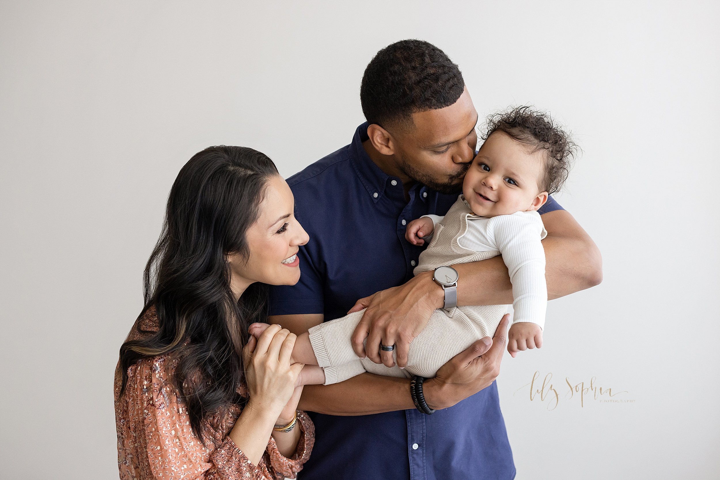  Family photograph of a father holding his seven month old son and giving him a kiss on the cheek as his wife holds her son’s feet and smiling while watching her husband enjoy their son taken in natural light in a studio near Virginia Highlands in At