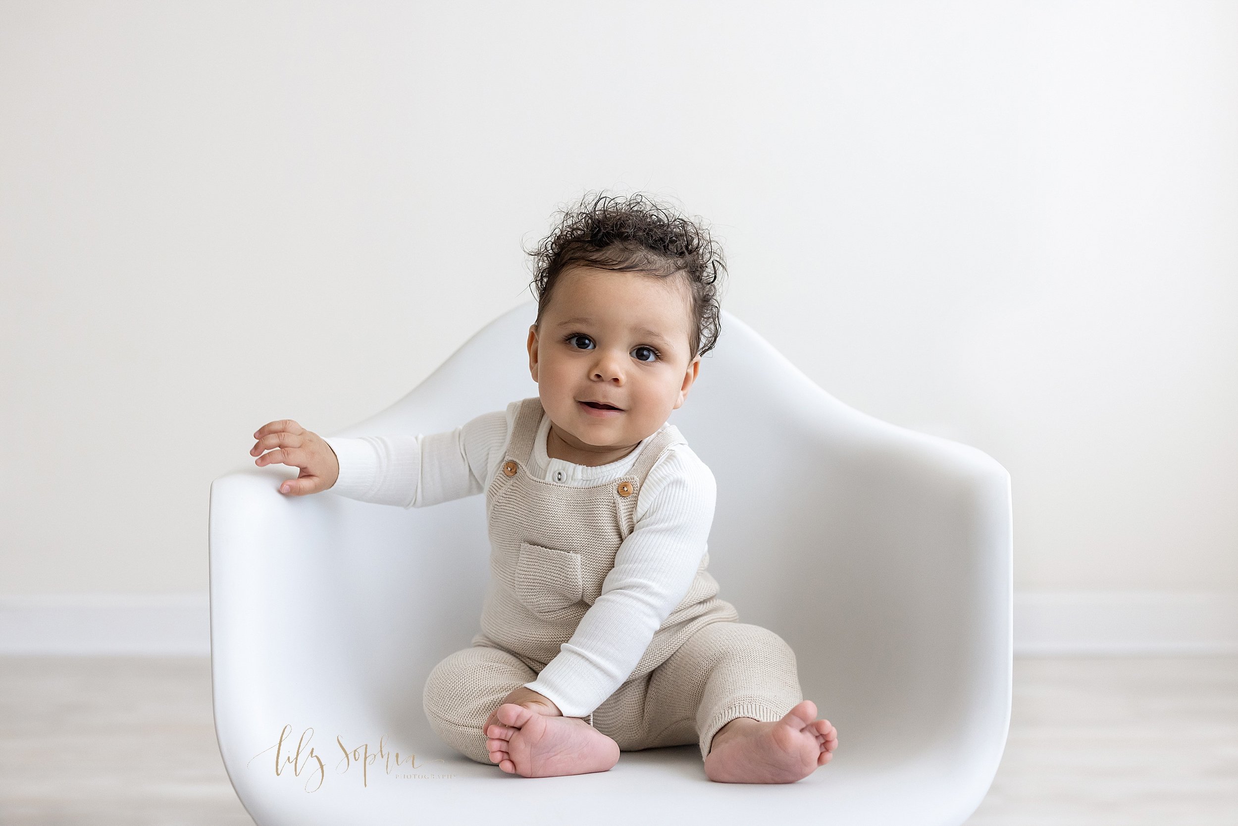  Milestone photo session of a seven month old little boy as he sits in a molded white chair with his right hand on the arm of the chair and his left hand touching his right ankle taken in a natural light studio near Oakhurst in Atlanta. 