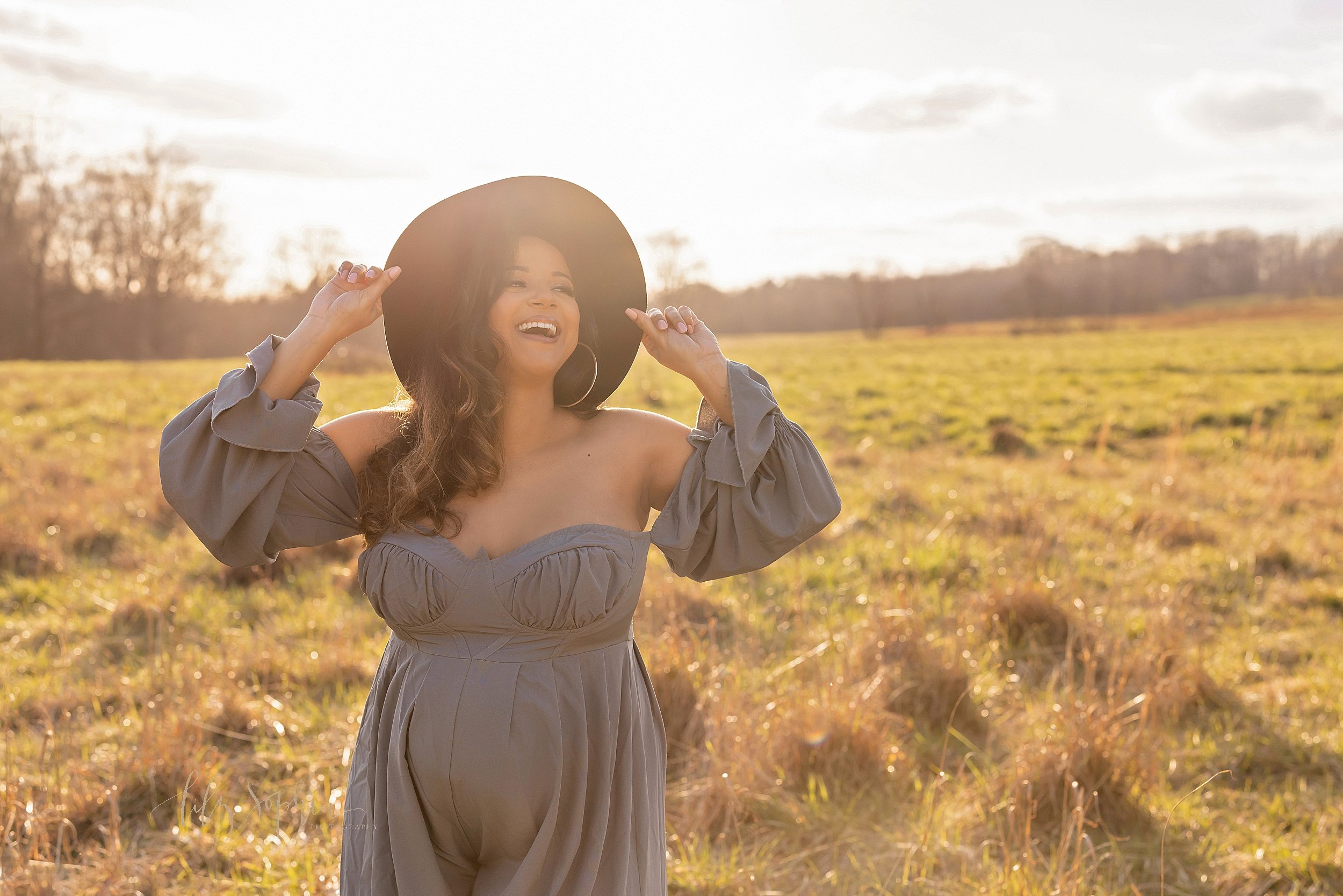  Maternity photo session of a pregnant mother wearing a wide-brimmed hat that she is holding with her hands as she stands at sunset in a field near Atlanta, Georgia. 