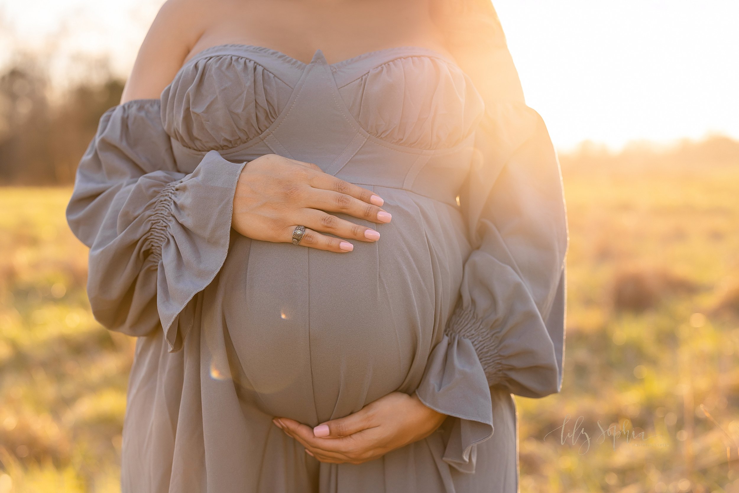  Close-up maternity picture of an expectant woman’s belly being framed by her hands as she stands in an Atlanta field at sunset. 