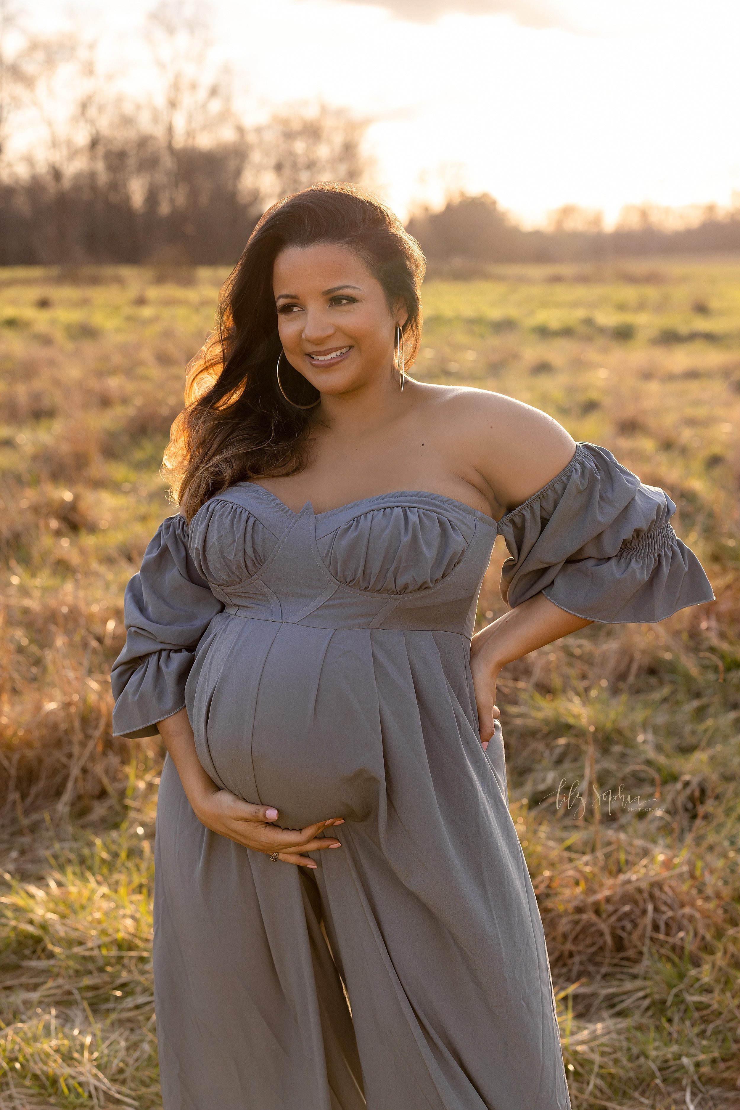  Maternity portrait of an expectant mother with her long brown hair flowing over her right shoulder as she stands at sunset in a field near Atlanta with her left hand on her hip and her right hand framing her belly.  