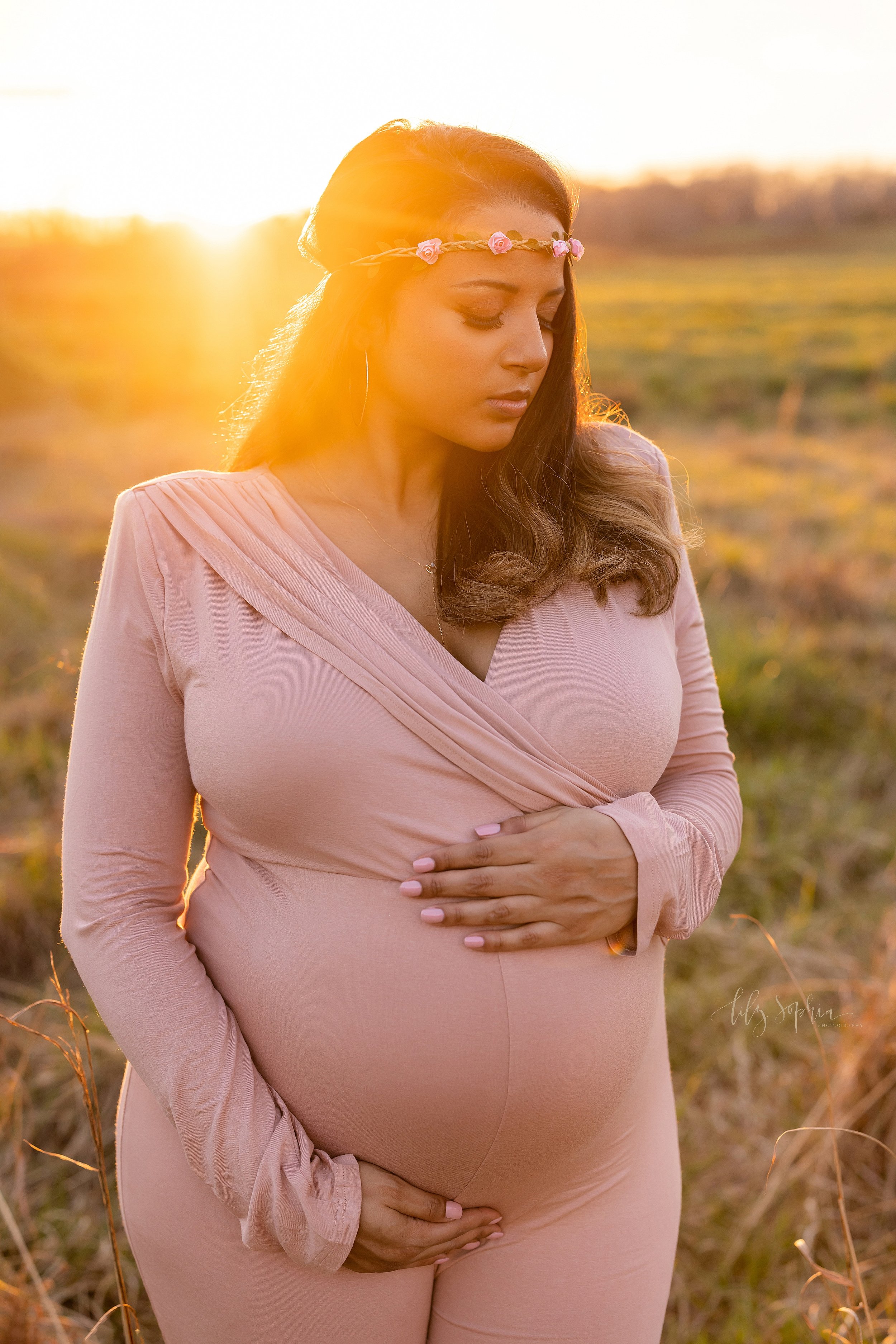  Maternity portrait of an expectant mother wearing a delicate rose band around her forehead and wearing a jersey knit full-length pant suit as she stands at sunset in a field framing her belly with her hands and looks down over her left shoulder take