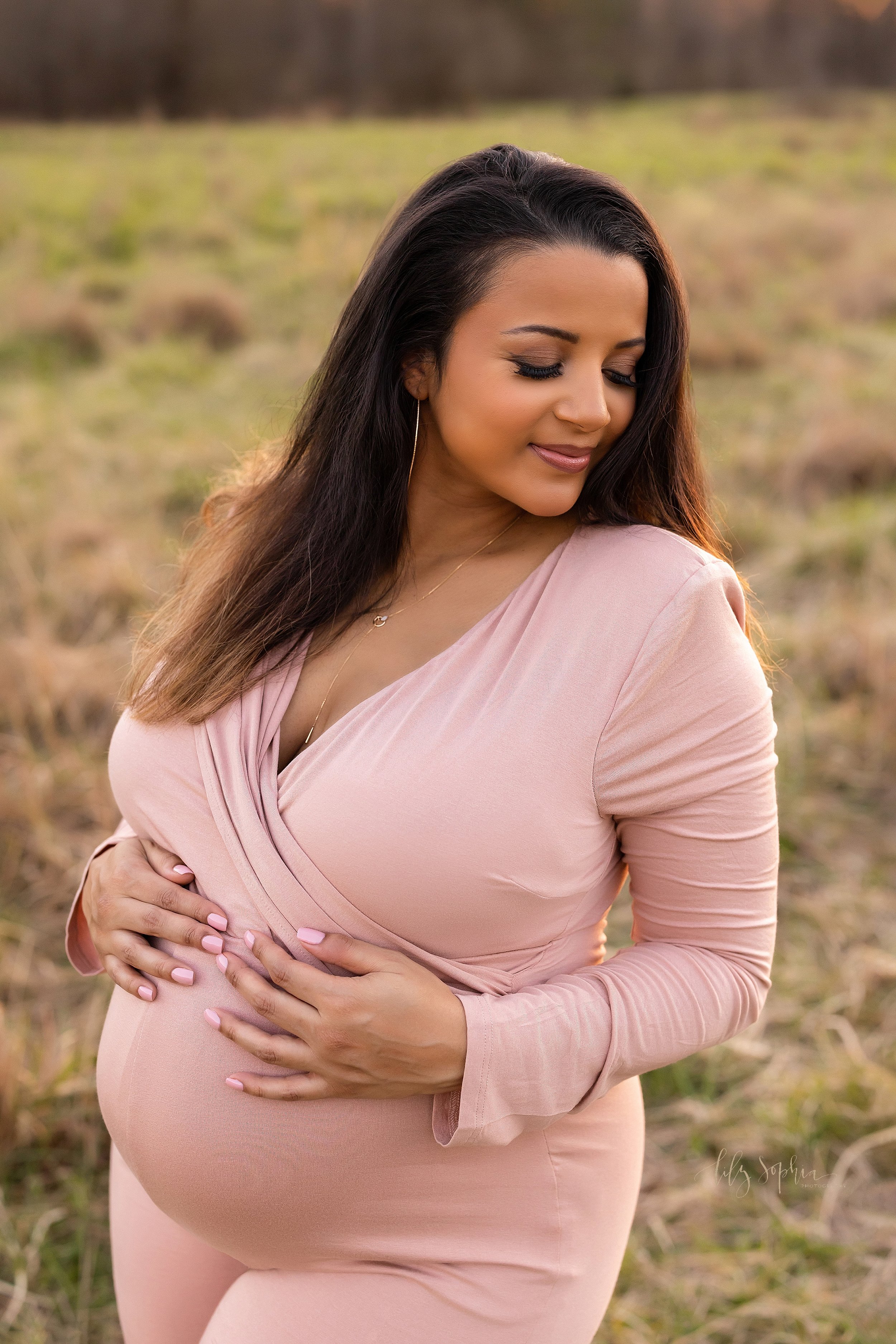  Maternity session with a pregnant woman wearing a pink jersey knit long-sleeved pant suit as she looks over her left shoulder while resting her hands on her belly and standing in a field near Atlanta at sunset. 