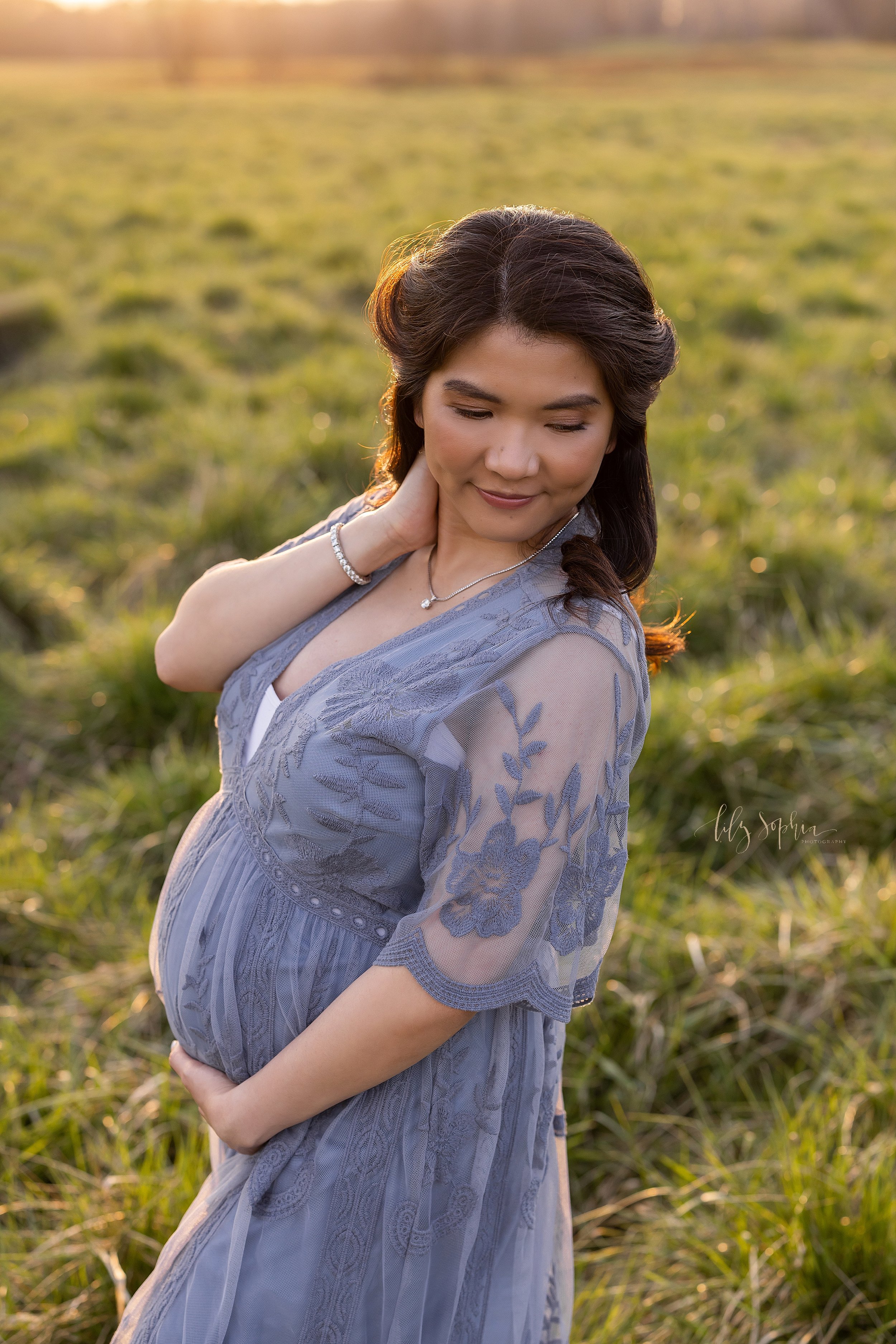  Maternity portrait of a pregnant mother as she places her right hand on her neck and looks over her left shoulder while holding her belly with her left hand as she stands at sunset in a field near Atlanta. 