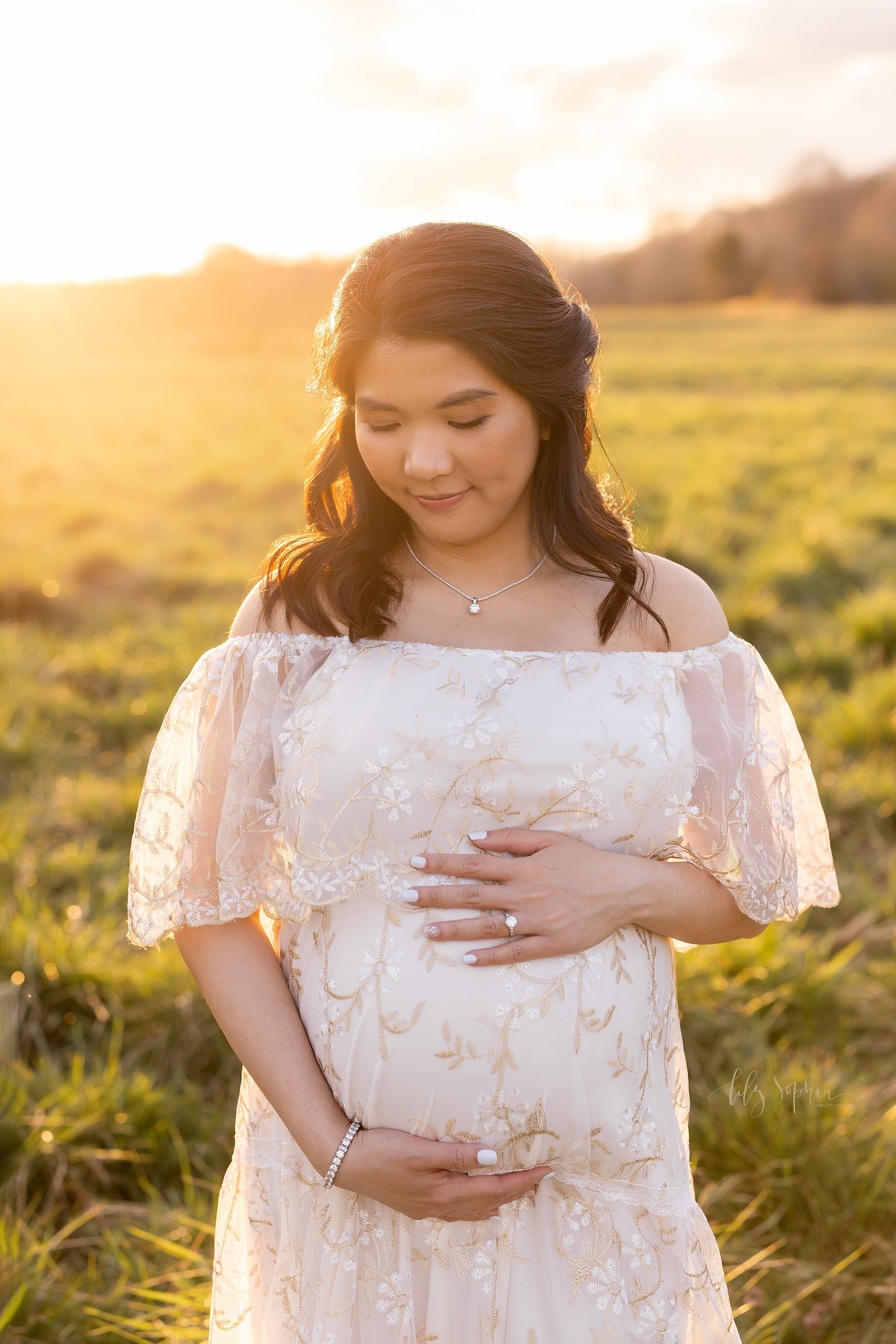 Maternity photo session of an Asian mother wearing an off the shoulder full-length gown with a wide lace bodice placing her hands to frame her belly as she contemplates the birth of her child with the sun setting behind her while standing in an Atla