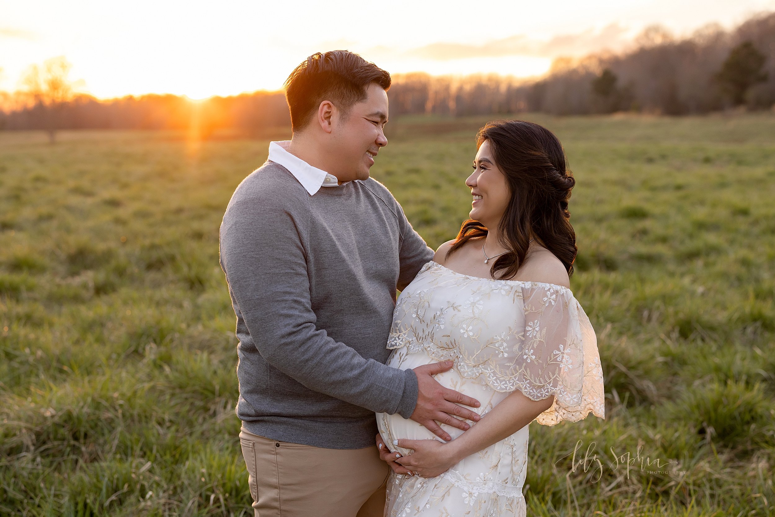  Maternity portrait of a husband and wife as they stand facing each other with the dad placing his hand on the child in utero and the mom frames her belly taken in an Atlanta field at sunset. 