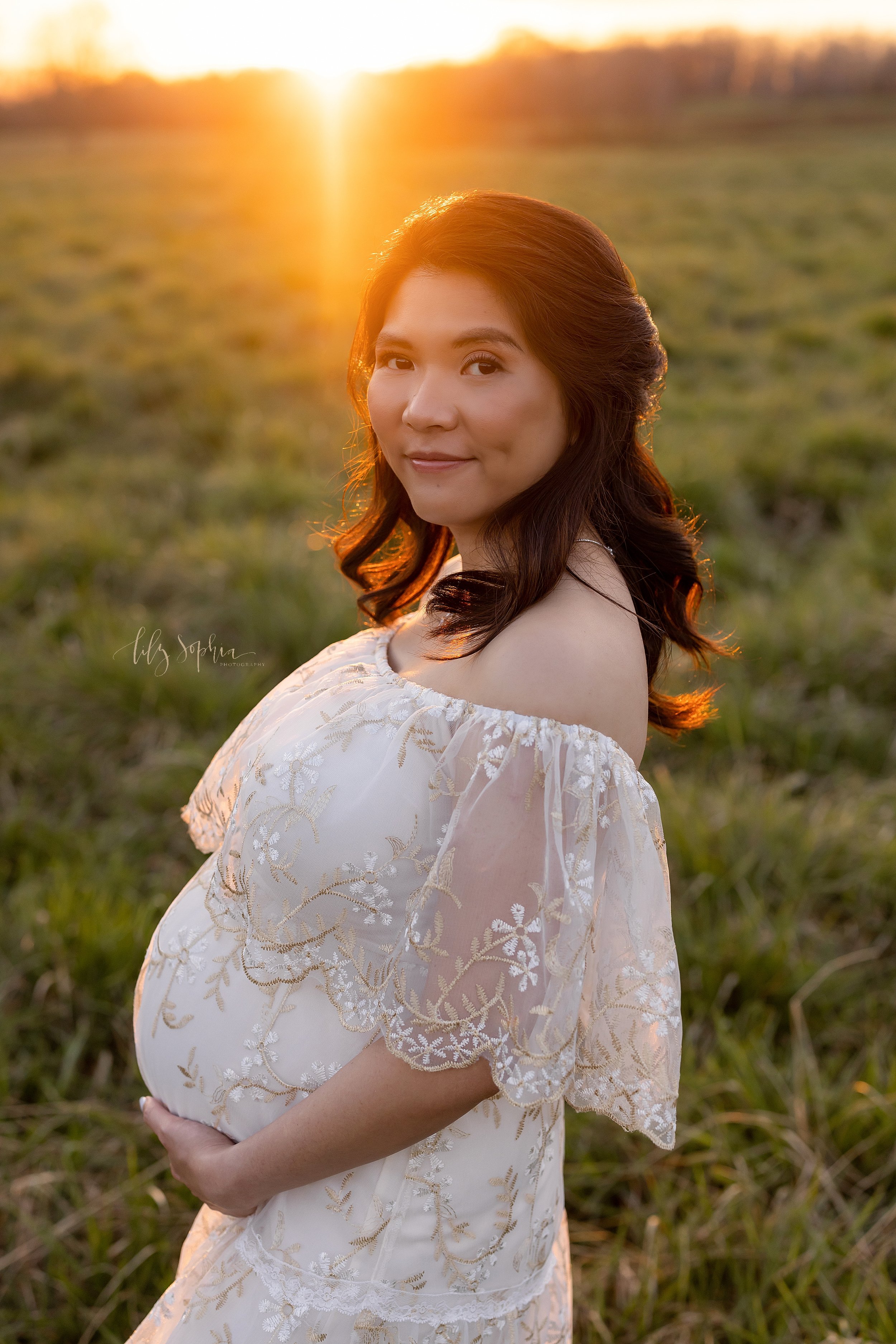  Maternity portrait of an Asian mother as she stands framing her belly while looking over her right shoulder as the sun sets in an Atlanta field. 