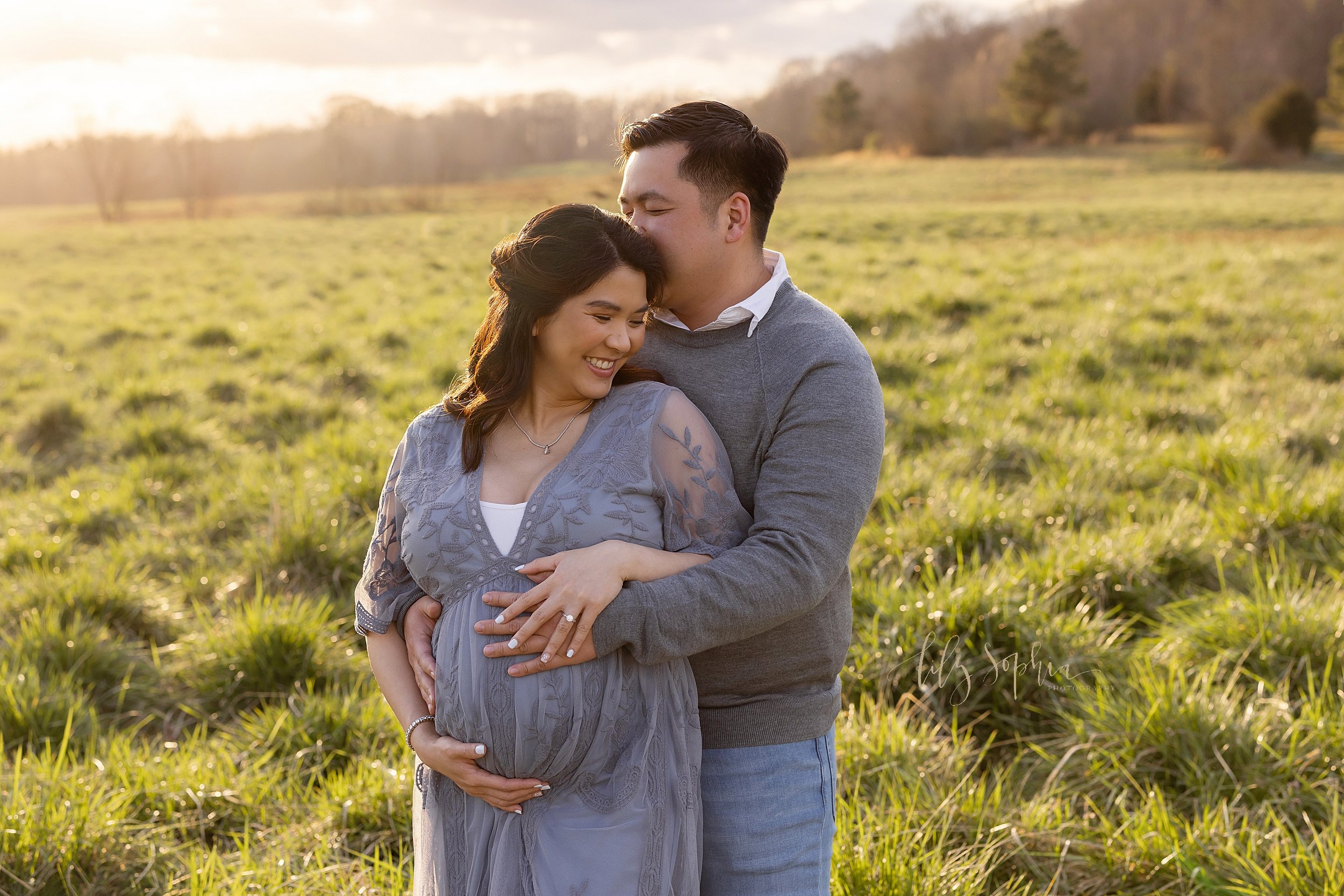  Maternity photo shoot of an expectant mother with her husband wrapping his arms around her from behind as both parents place their hands on their child in utero and the husband kisses his wife’s head while they stand at sunset in a field near Atlant
