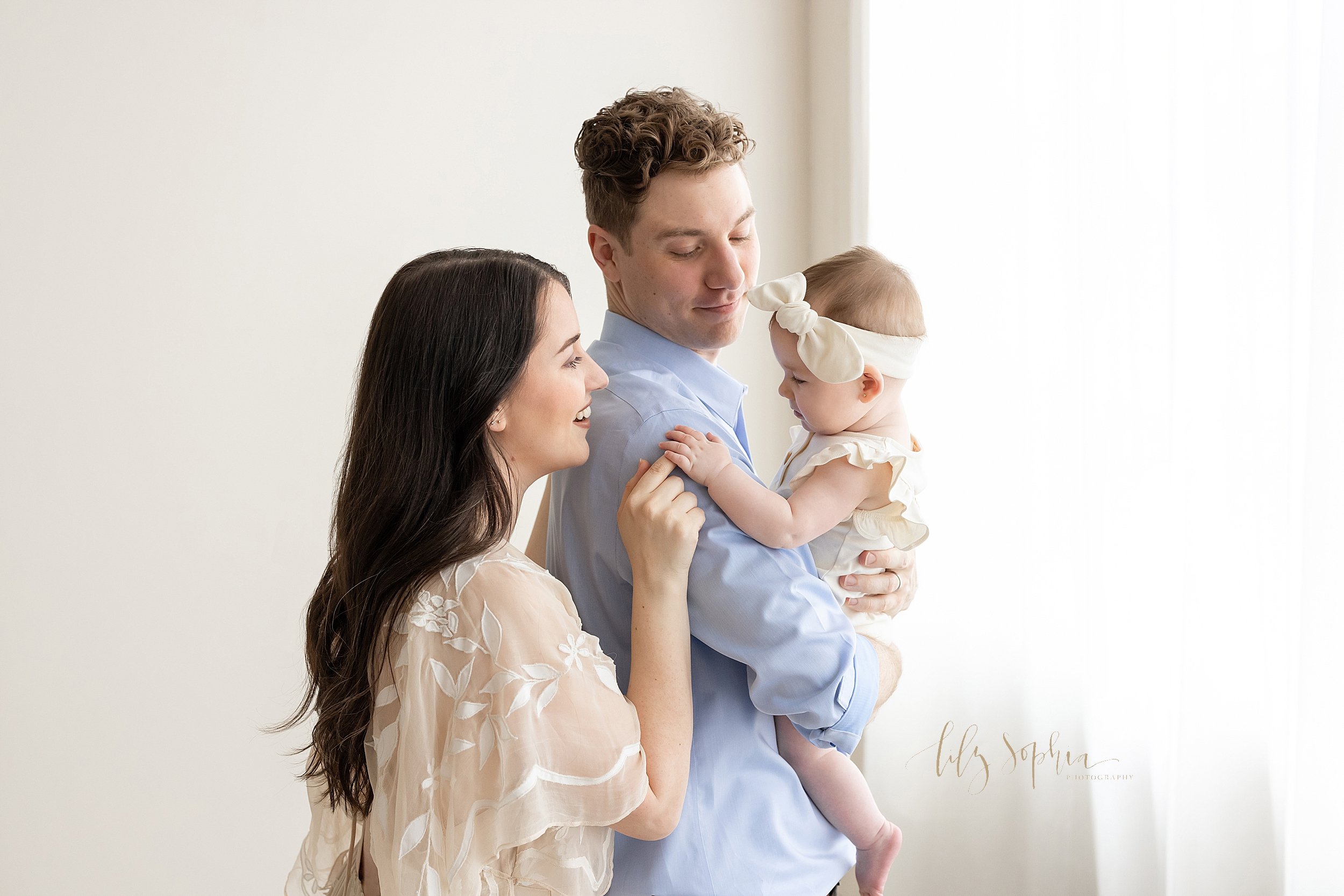  Family photograph of a father holding his six month old baby girl in his arms as his wife stands behind her and their daughter plays with her finger taken in natural light in a studio near Buckhead in Atlanta, Georgia. 