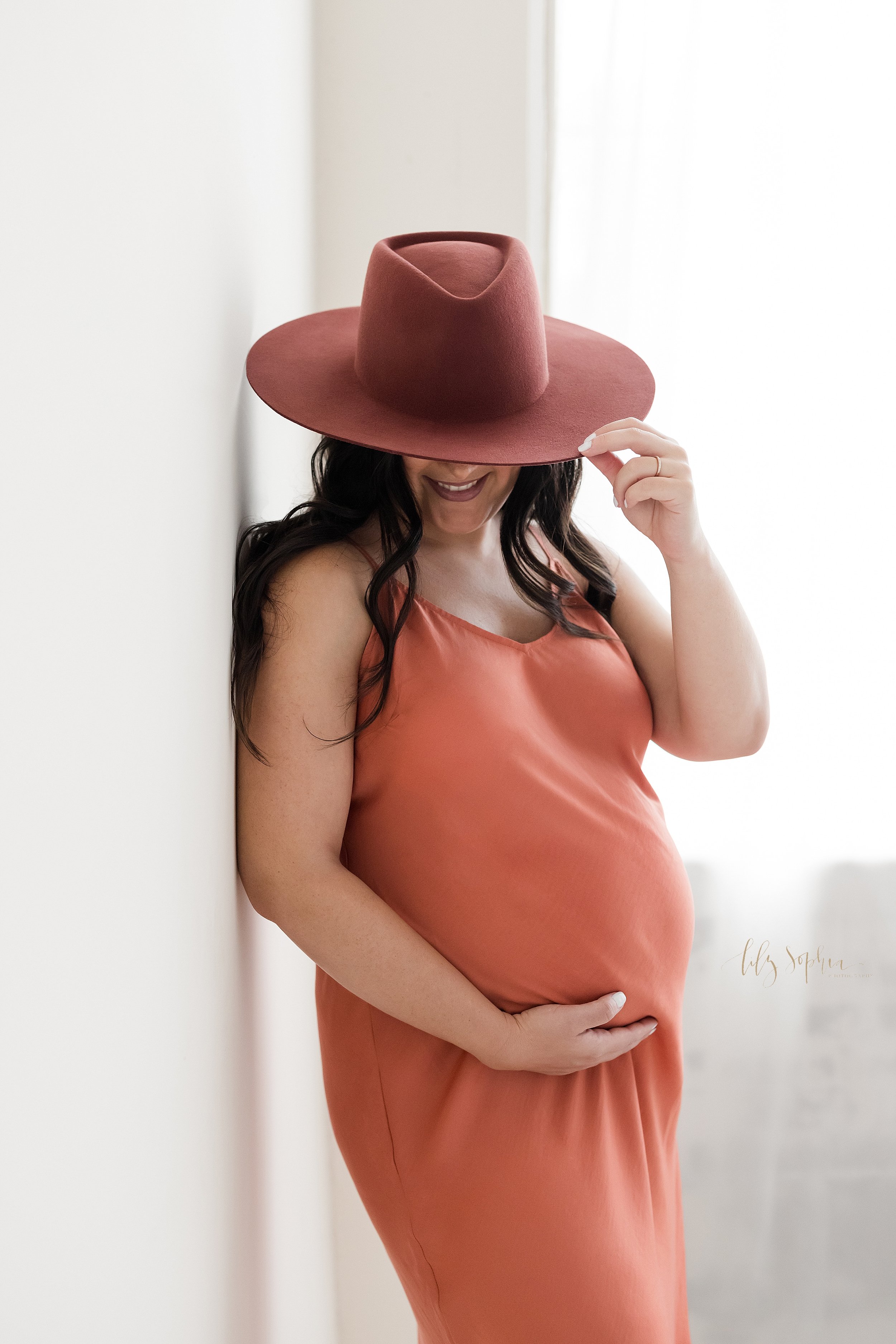  Maternity portrait of an expectant mother as she stands wearing a wide brimmed hat and a spaghetti strapped full-length jersey gown with her right shoulder against a wall and her right hand holding her belly and her left hand holding the brim of the