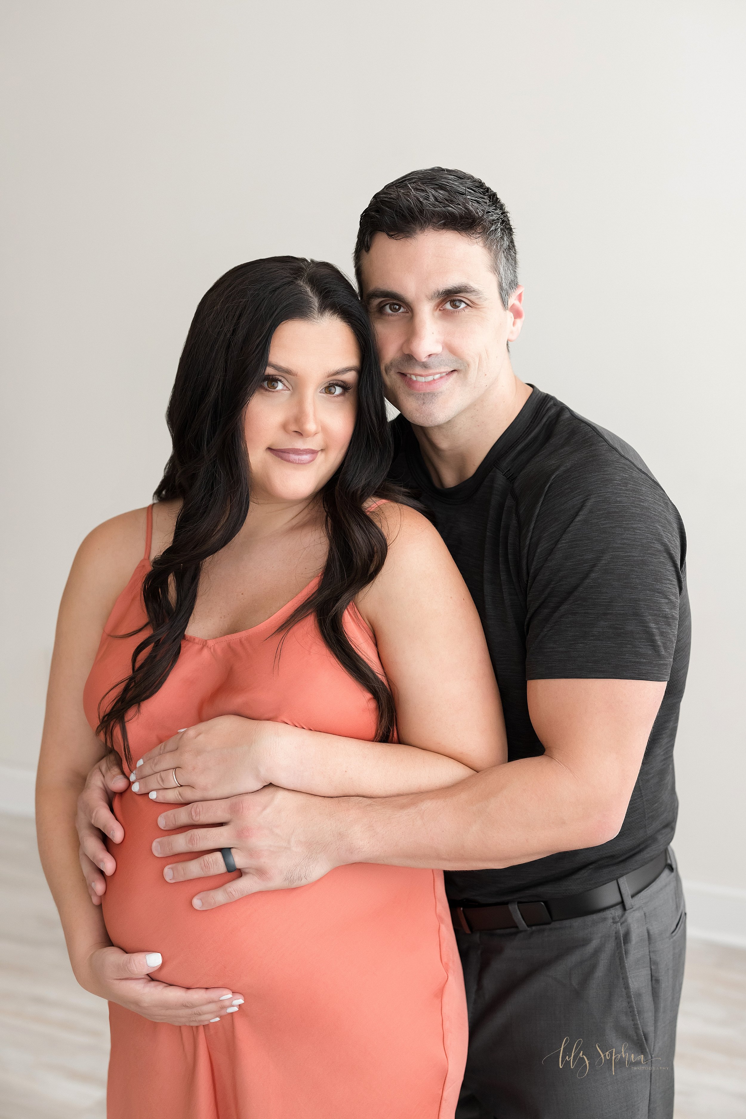  Maternity photograph of an expectant couple as the husband wraps his arms around his wife from behind and both of them hold their child in utero taken in a studio near Oakhurst in Atlanta that uses natural lighting. 