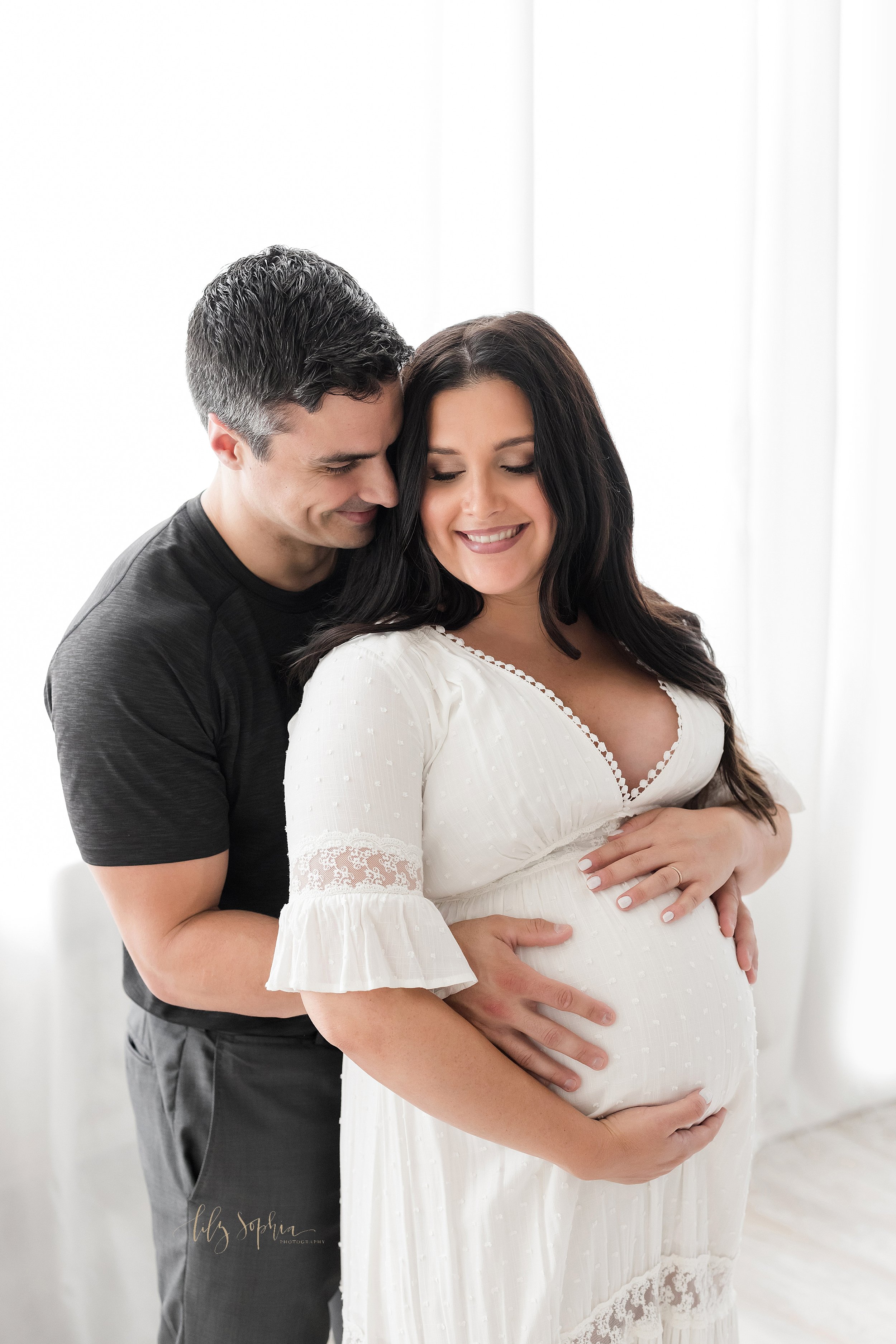  Maternity photo session of a pregnant mother standing with her husband behind her as she frames her belly with her hands and he holds his child in utero in a studio in front of a window streaming natural light near Poncey Highlands in Atlanta, Georg