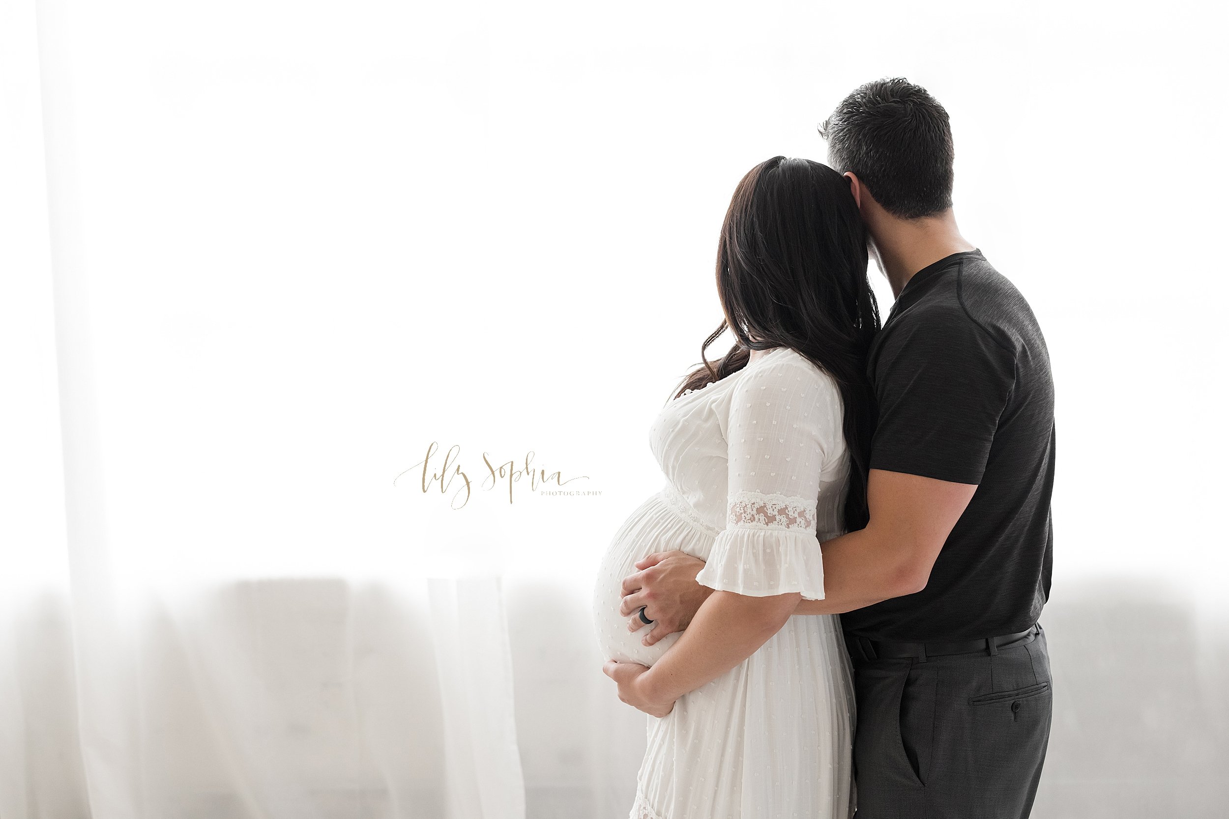  Maternity photo of a husband standing behind his pregnant wife with his arms wrapped around her with his hands resting on their child in utero and his wife with her hands holding her belly as the two of them look out a natural light window in a stud