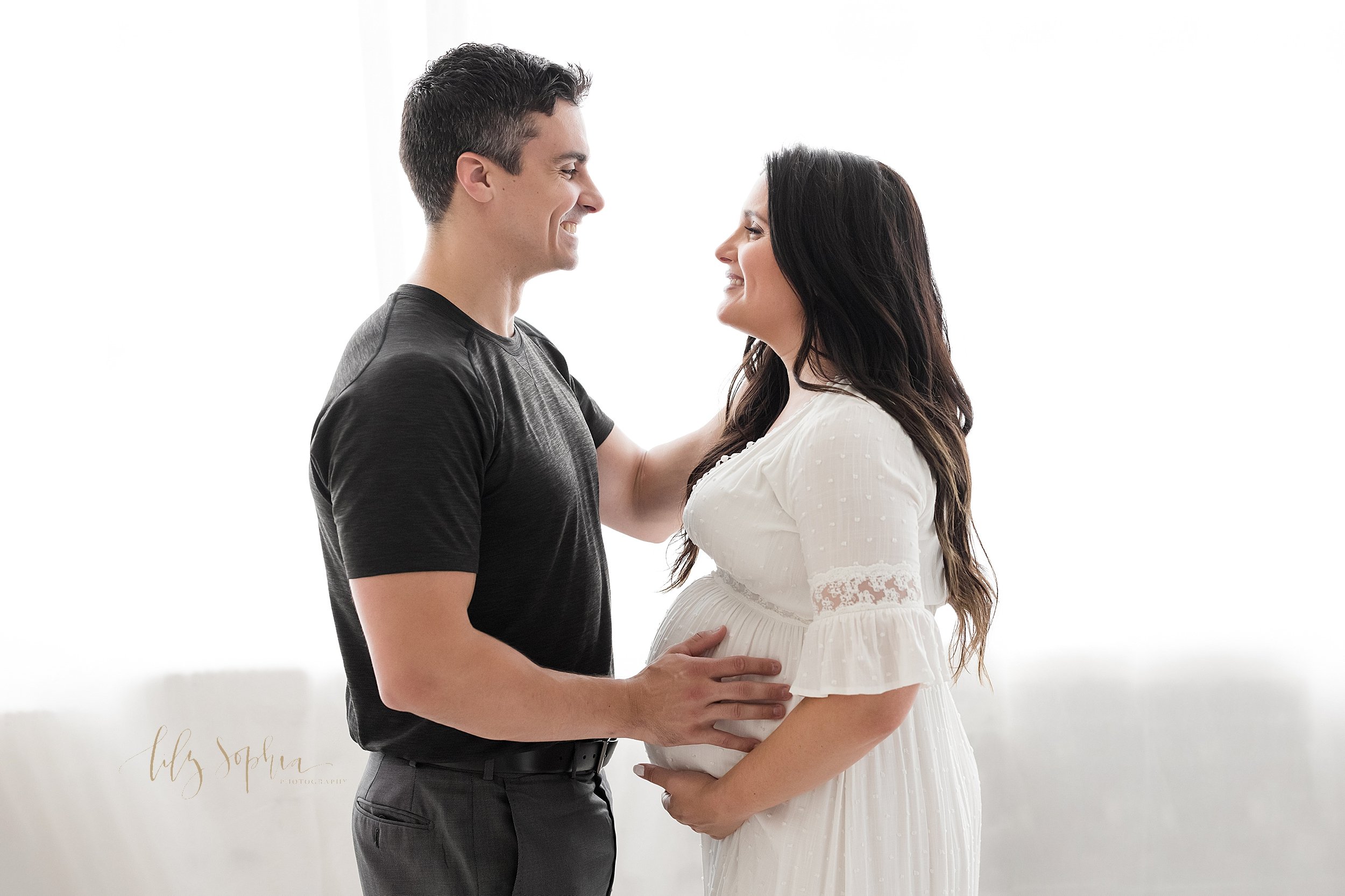  Maternity photo shoot of an expectant couple with the pregnant mother holding the base of her belly as her husband faces her and places his right hand on their child in utero while they stand together in front of a natural light window in a studio i