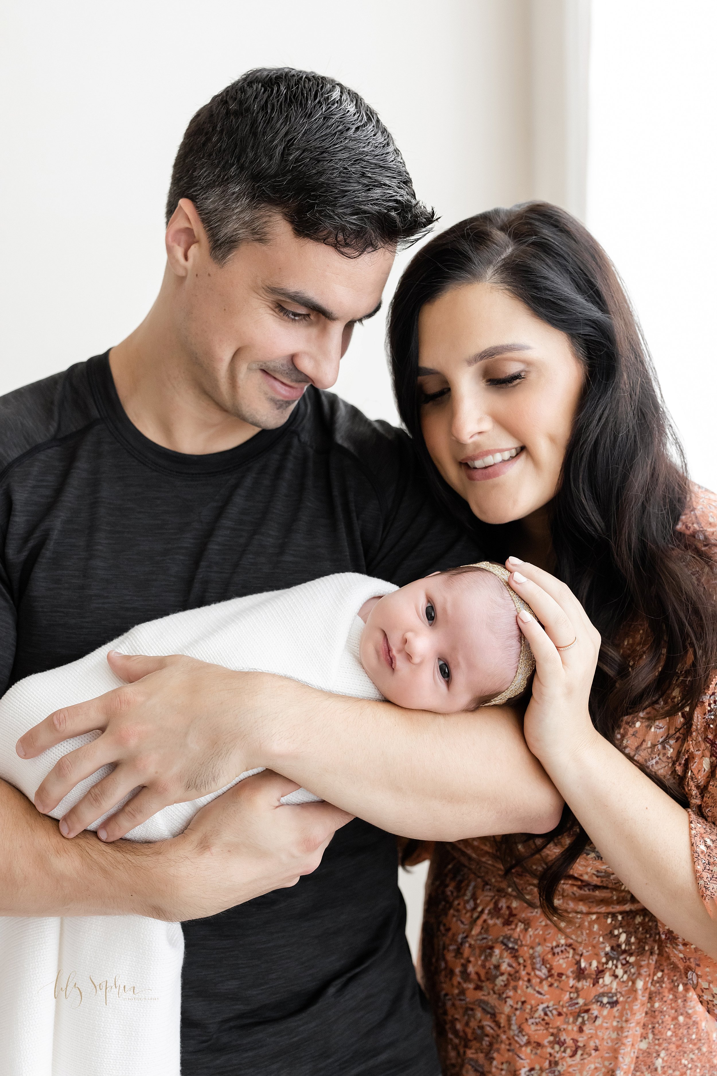  Family newborn portrait of a father holding his awake newborn baby girl in his arms as his wife stands to his right side and places her hand on her daughter’s head taken in front of a window streaming natural light in a studio near Ansley Park in At