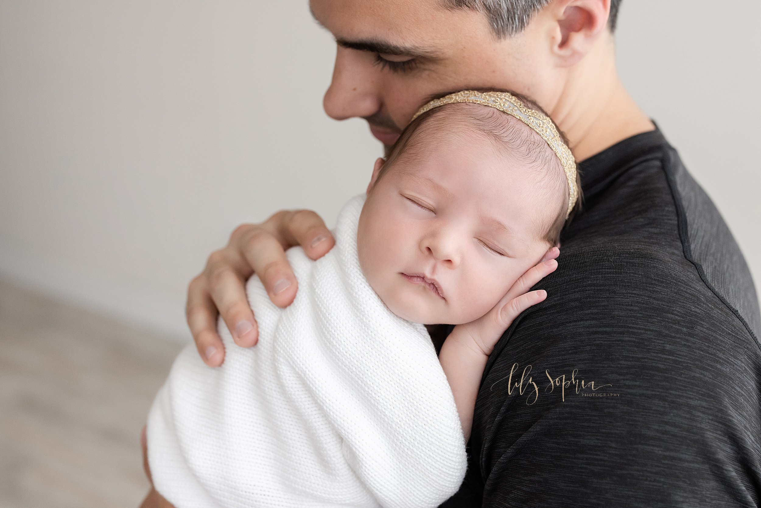  Close-up newborn portrait of a contentedly sleeping baby girl as her father holds her against his right shoulder taken in a studio near Buckhead in Atlanta that uses natural light. 