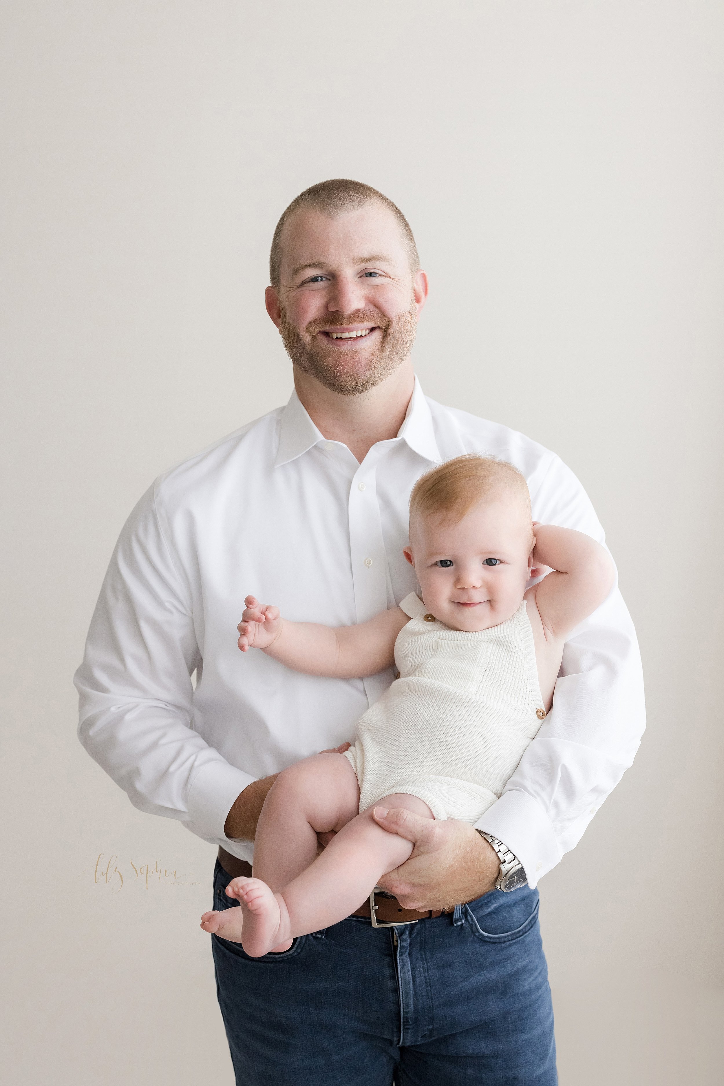  Family picture of a father standing in a studio in front of a window streaming natural light and holding his six month old baby boy in front of him taken near Kirkwood in Atlanta, Georgia. 