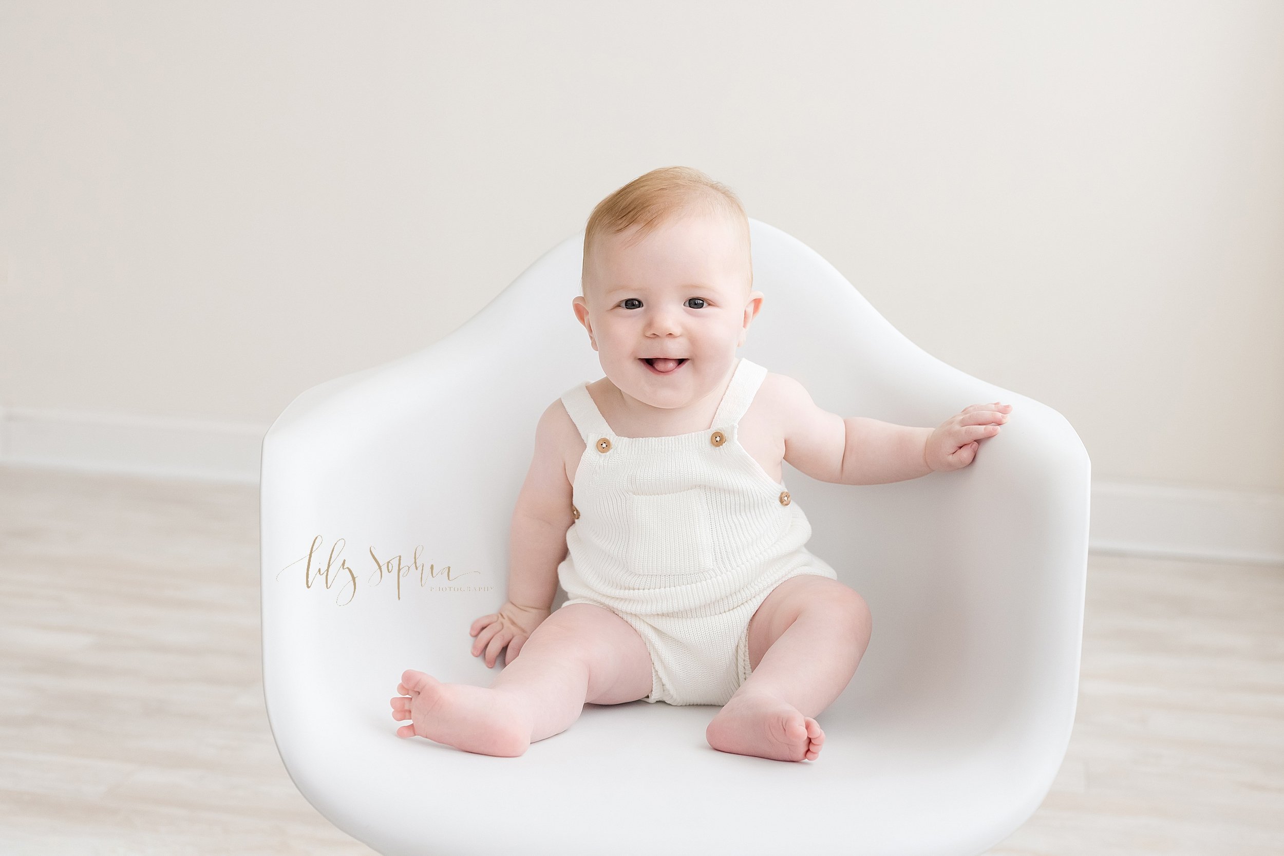  Milestone photo of a red-headed six month old baby boy sitting in a white molded chair in front of a window streaming natural light and sticking out his tongue taken in a studio near Decatur in Atlanta. 