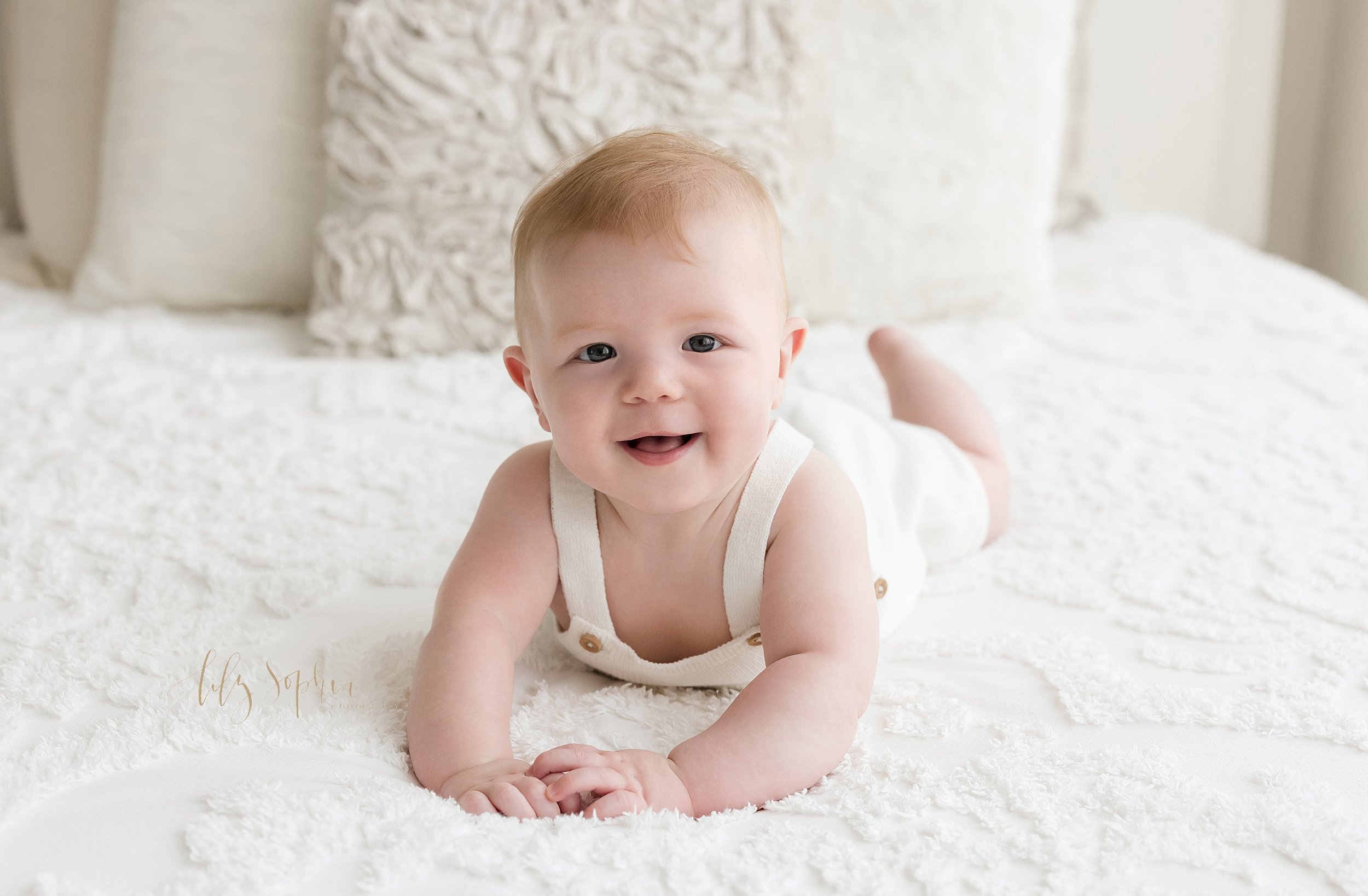  Milestone photograph of a smiling six month old baby boy as he lies on his stomach on a bed in a studio near Midtown in Atlanta that uses natural lighting. 