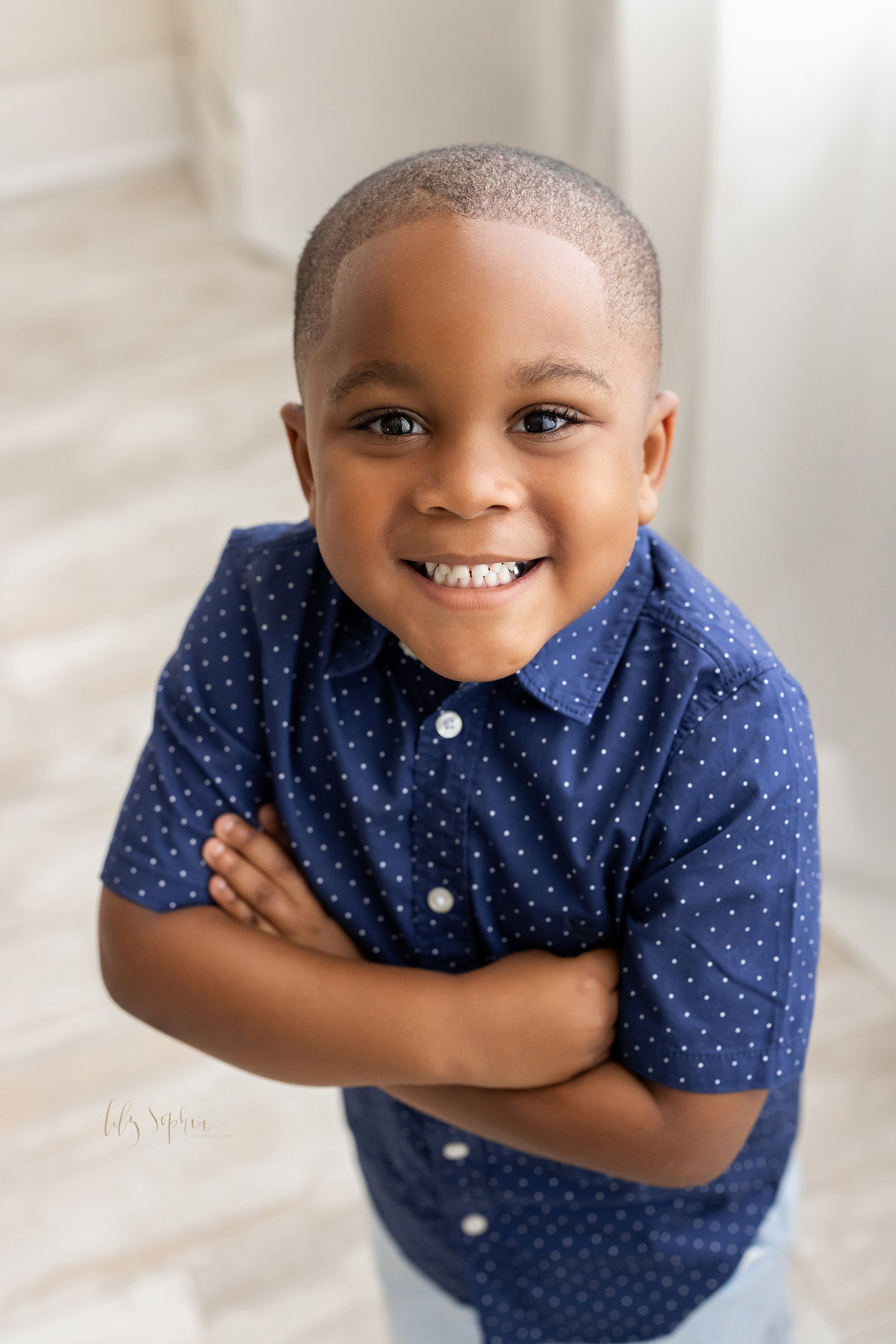  Close-up overhead photograph of a five year old African-American boy standing in front of a natural light window smiling with his arms crossed against his chest taken in a studio near Virginia Highlands in Atlanta. 