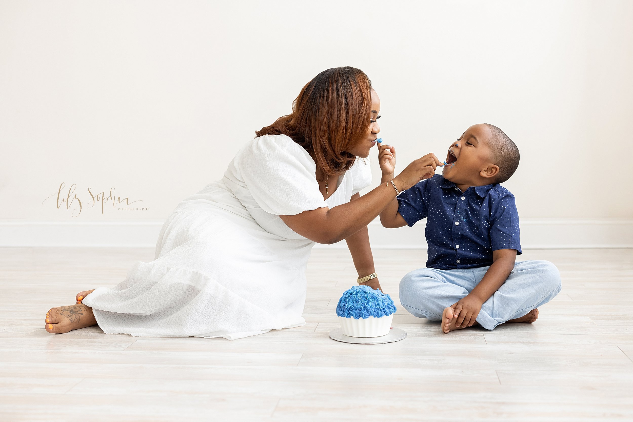  Milestone photograph of an African-American mother sitting on the wooden floor next to her five year old son as the two of them give tastes of icing to one another from the large blue iced cupcake taken near Decatur in Atlanta in a natural light stu